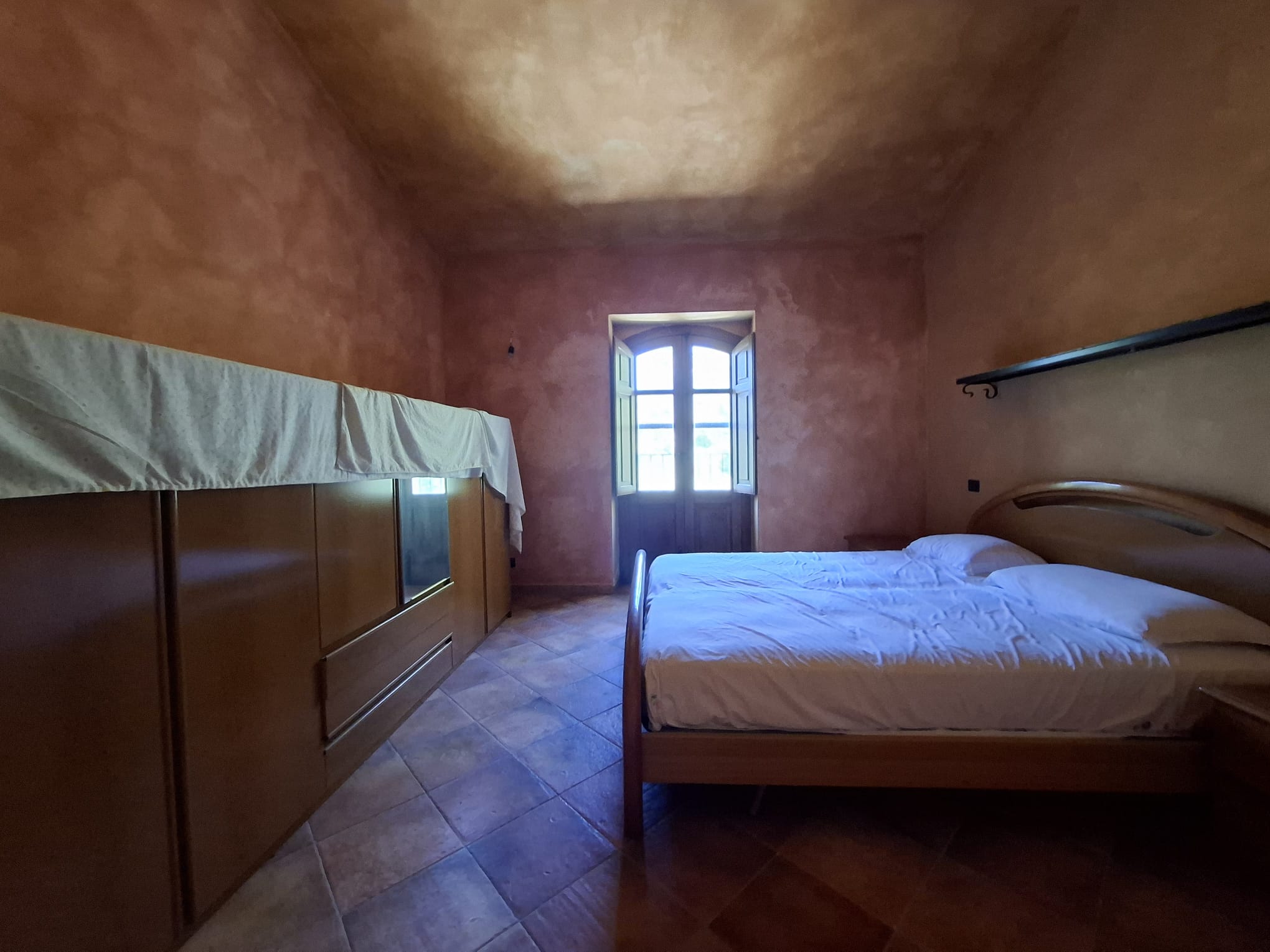Riproduzione di una camera da letto con pareti color terracotta, grande finestra che permette l'ingresso di luce naturale, letto matrimoniale e armadio in legno, ambiente accogliente e rustico.