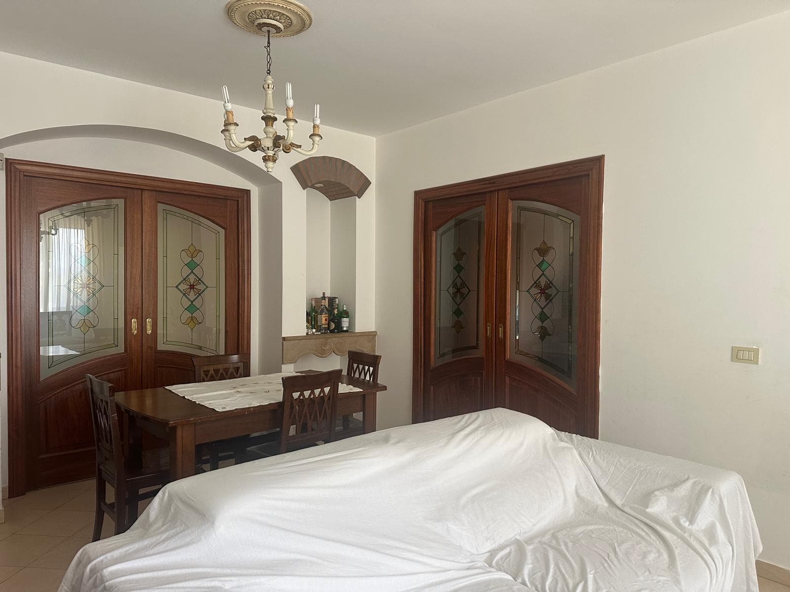 Interno di un appartamento con pareti bianche, tavolo in legno, sedie, e porte in legno con vetri decorativi, illuminato da lampadario classico.