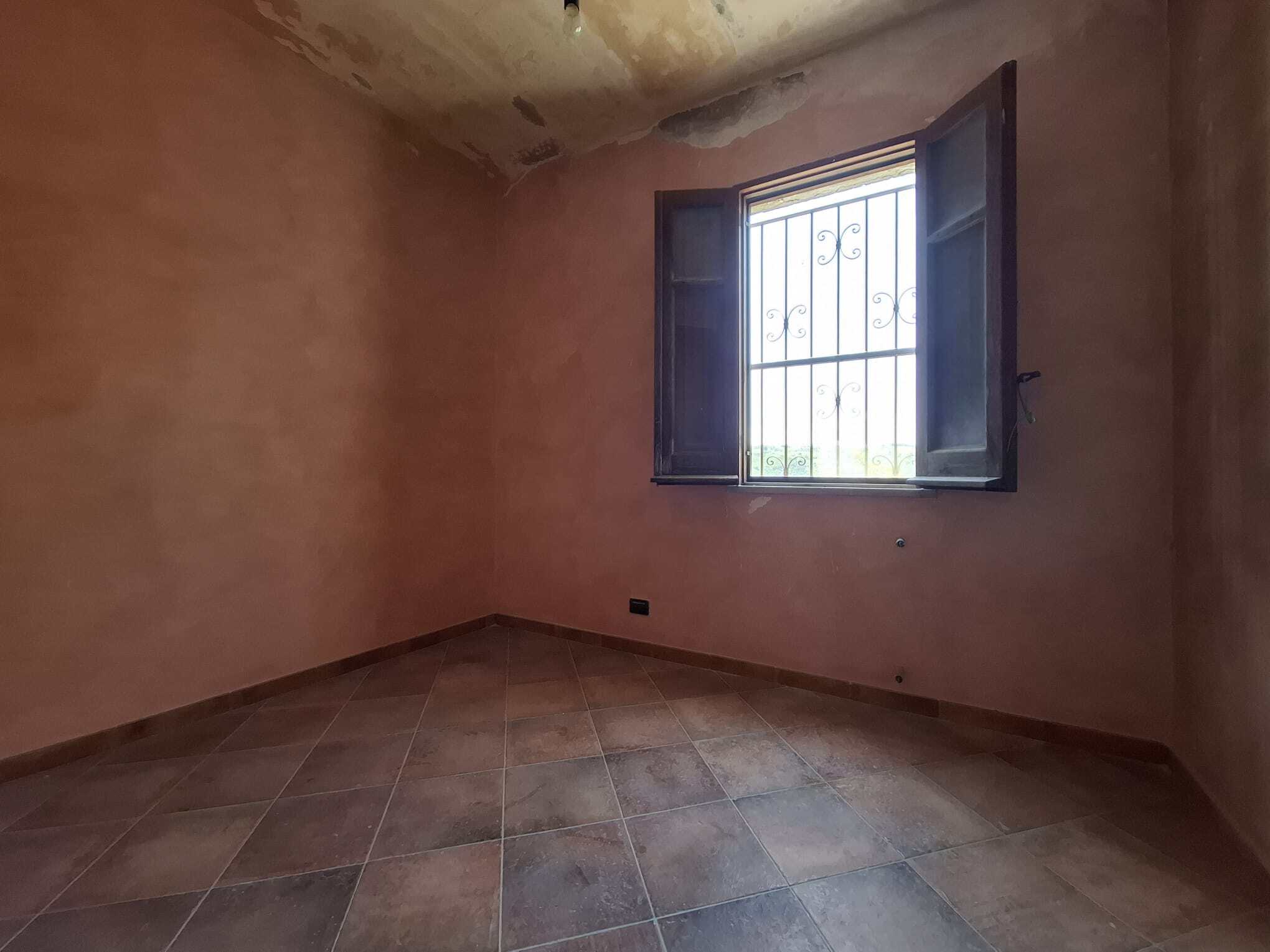 Ampia stanza vuota con pareti in tonalità calde, finestra con grate in ferro e persiane aperte, pavimento in piastrelle di ceramica, luce naturale che entra dalla finestra, ideale per ristrutturazione.