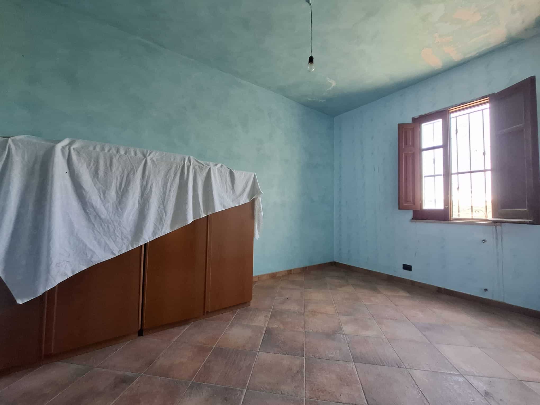Stanza vuota con pareti azzurre, finestra aperta e armadio coperto da un telo bianco, ambiente luminoso e spazioso.