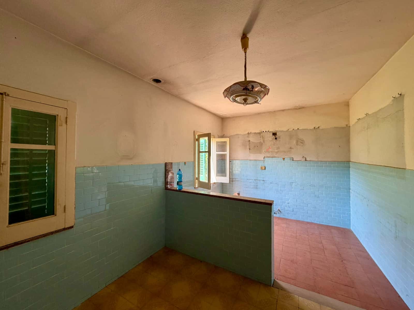 Interno di una stanza abitabile con pareti azzurre, finestra con persiane verdi e soffitto con lampadario, ambiente luminoso e spazioso.