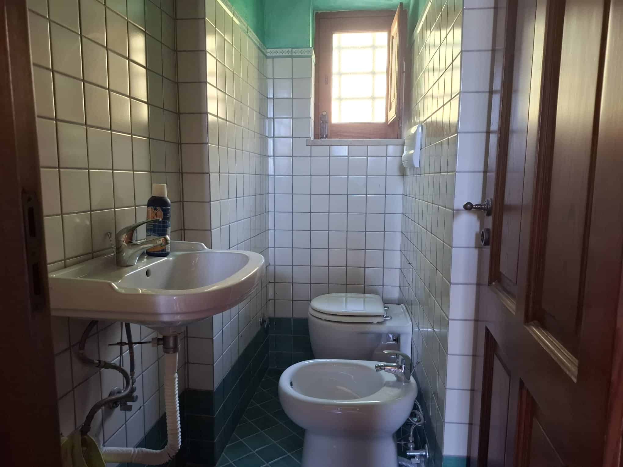 Lavandino e WC in bagno con pareti rivestite di piastrelle bianche e finestra con luce naturale, ambiente pulito e funzionale.