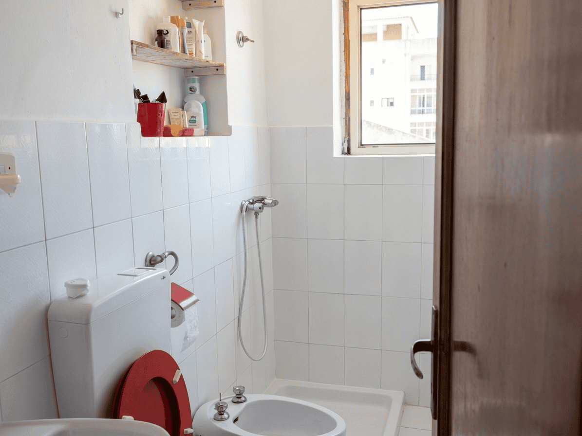 Lavabo e bidet in un bagno con pareti rivestite di piastrelle bianche, finestra che permette l'ingresso di luce naturale, e scaffale con prodotti per l'igiene personale.