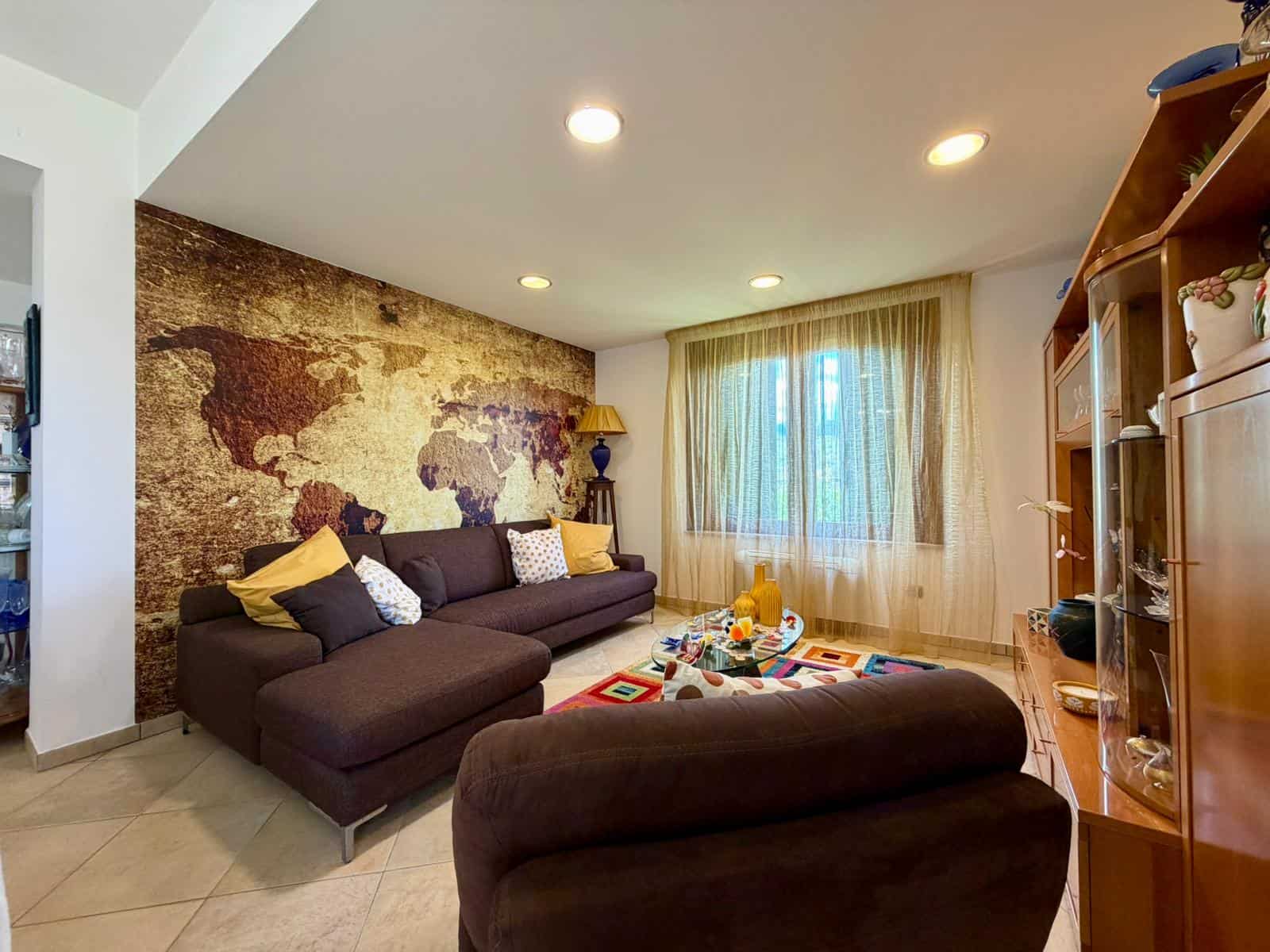 Interno soggiorno di una villa con parete decorativa con mappa del mondo, divano e poltrona, illuminazione soffusa e grande finestra con tende leggere.