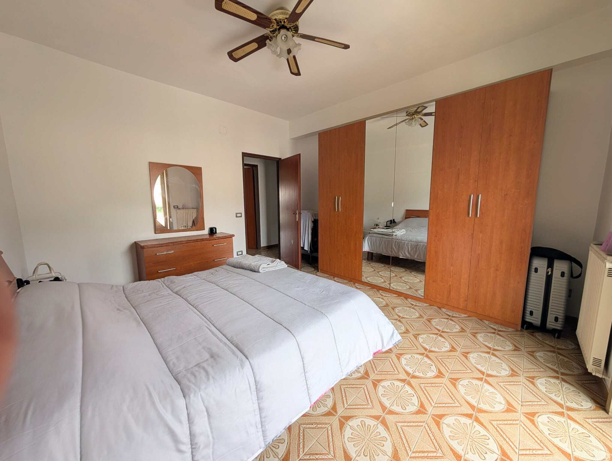 Camera da letto con letto matrimoniale, armadio a due ante con specchio e mobili in legno, pavimento in piastrelle decorative, ventilatore a soffitto, ambiente luminoso e ordinato.