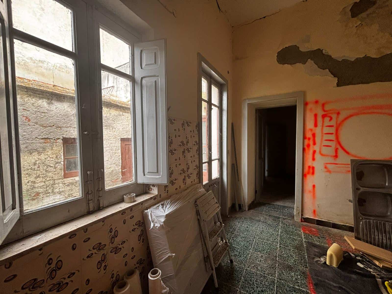 Interno di un appartamento in ristrutturazione con pareti danneggiate, finestre grandi e pavimento in piastrelle, pronto per lavori di ristrutturazione e riqualificazione.