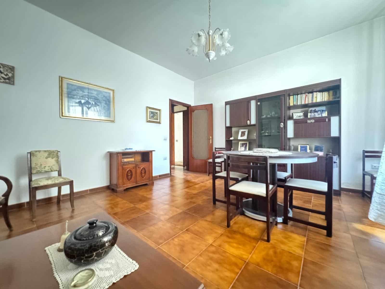 Panca in legno e pareti bianche in un soggiorno spazioso con pavimento in piastrelle di ceramica, arredamento classico, quadri alle pareti e grande libreria con libri e oggetti decorativi.