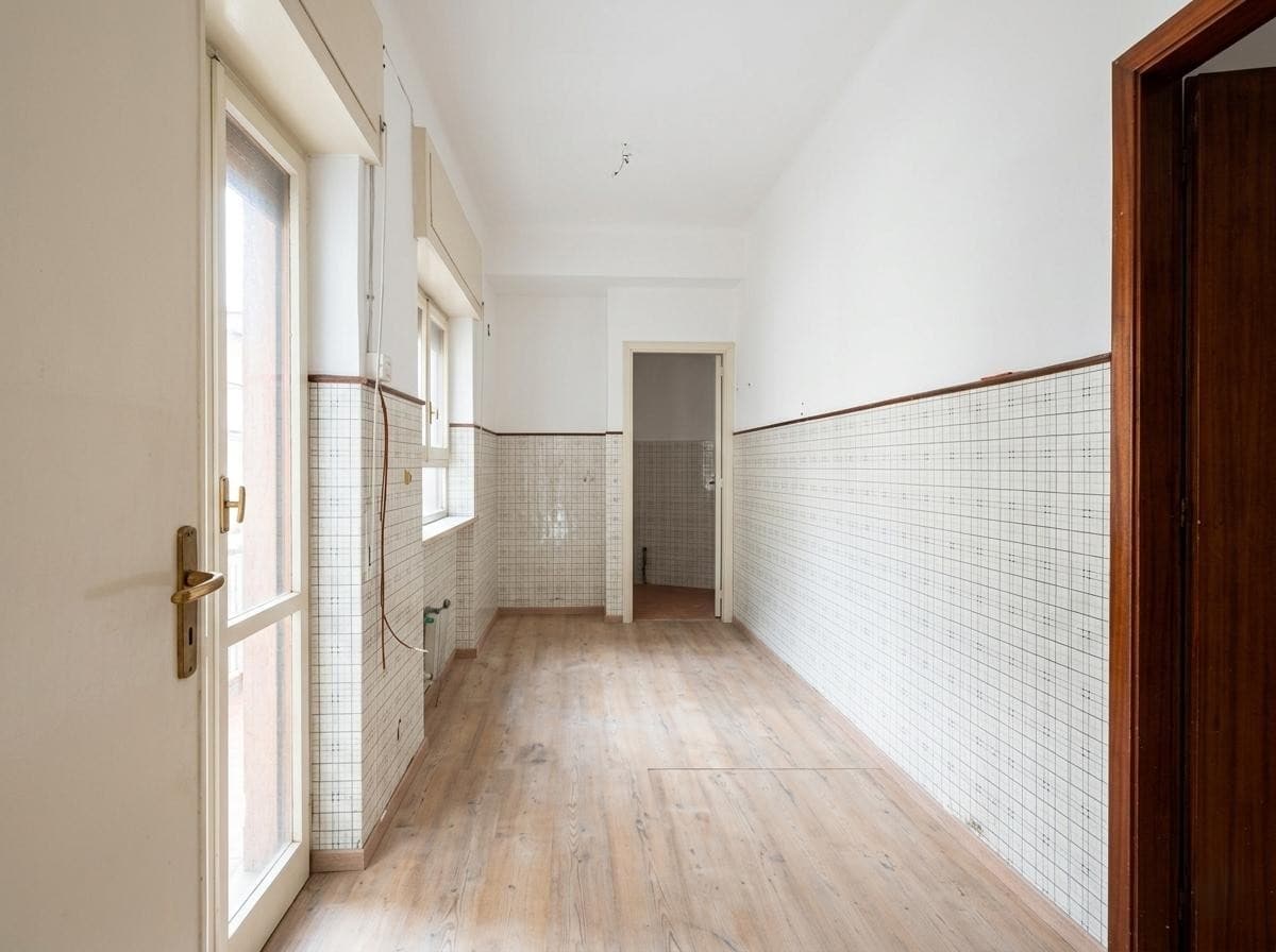 Ampio spazio vuoto in cucina con pareti rivestite di piastrelle bianche e pavimento in legno chiaro, illuminato da grandi finestre e porta finestra che permette l'accesso all'esterno.