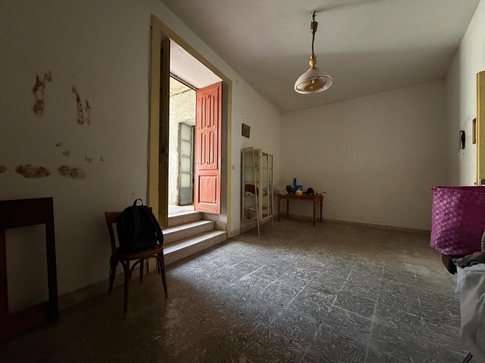 Interno di una casa indipendente con pareti danneggiate e pavimento in pietra, ingresso con porta rossa e pochi mobili.