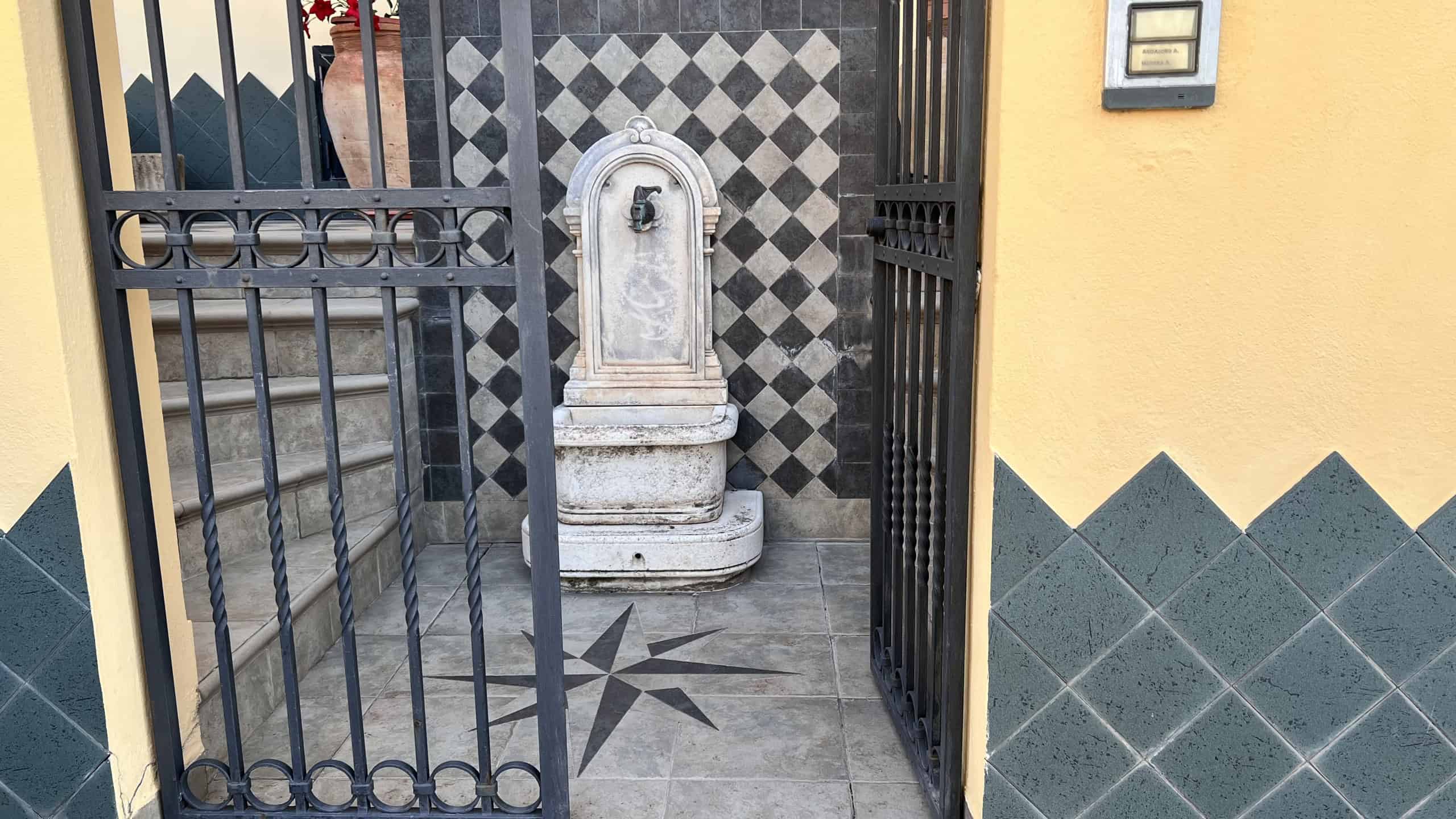 Fontana decorativa in cortile di una villa in vendita a Villafranca Tirrena, con dettagli in pietra e un cancello in ferro battuto.