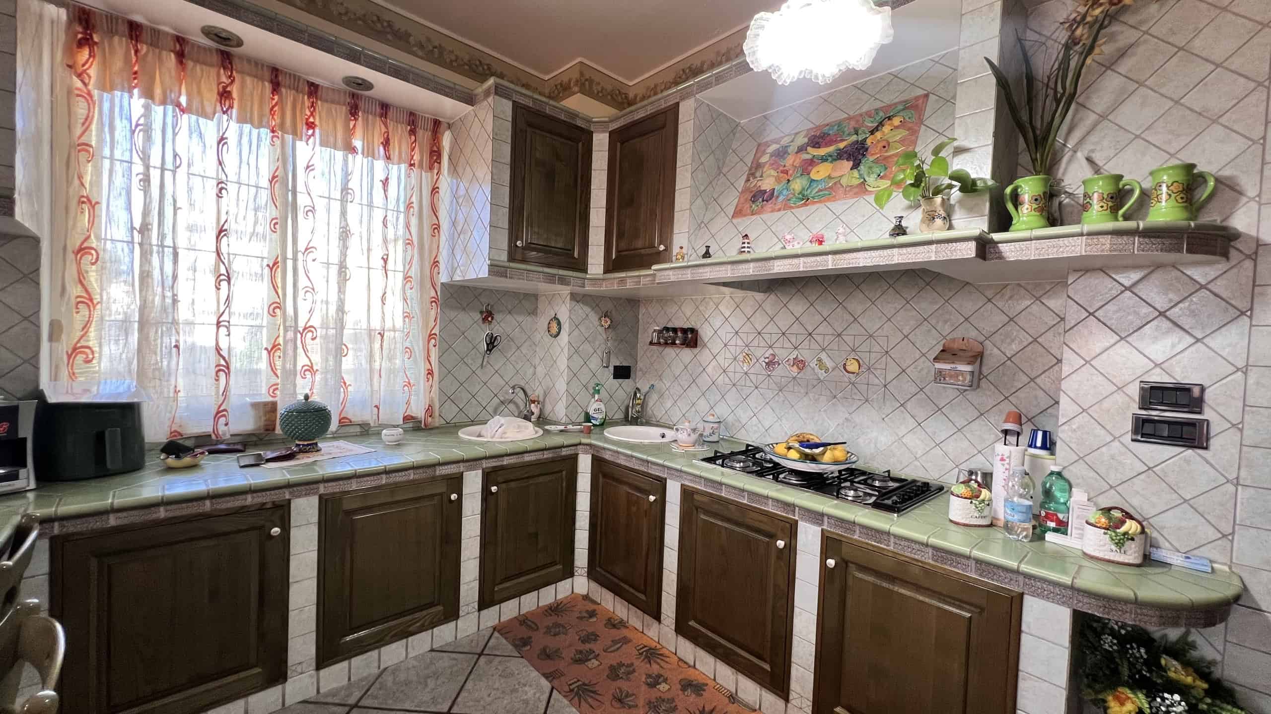 Cucina spaziosa con mobili in legno scuro, piastrelle beige e dettagli decorativi colorati, illuminata da luce naturale e lampadario.