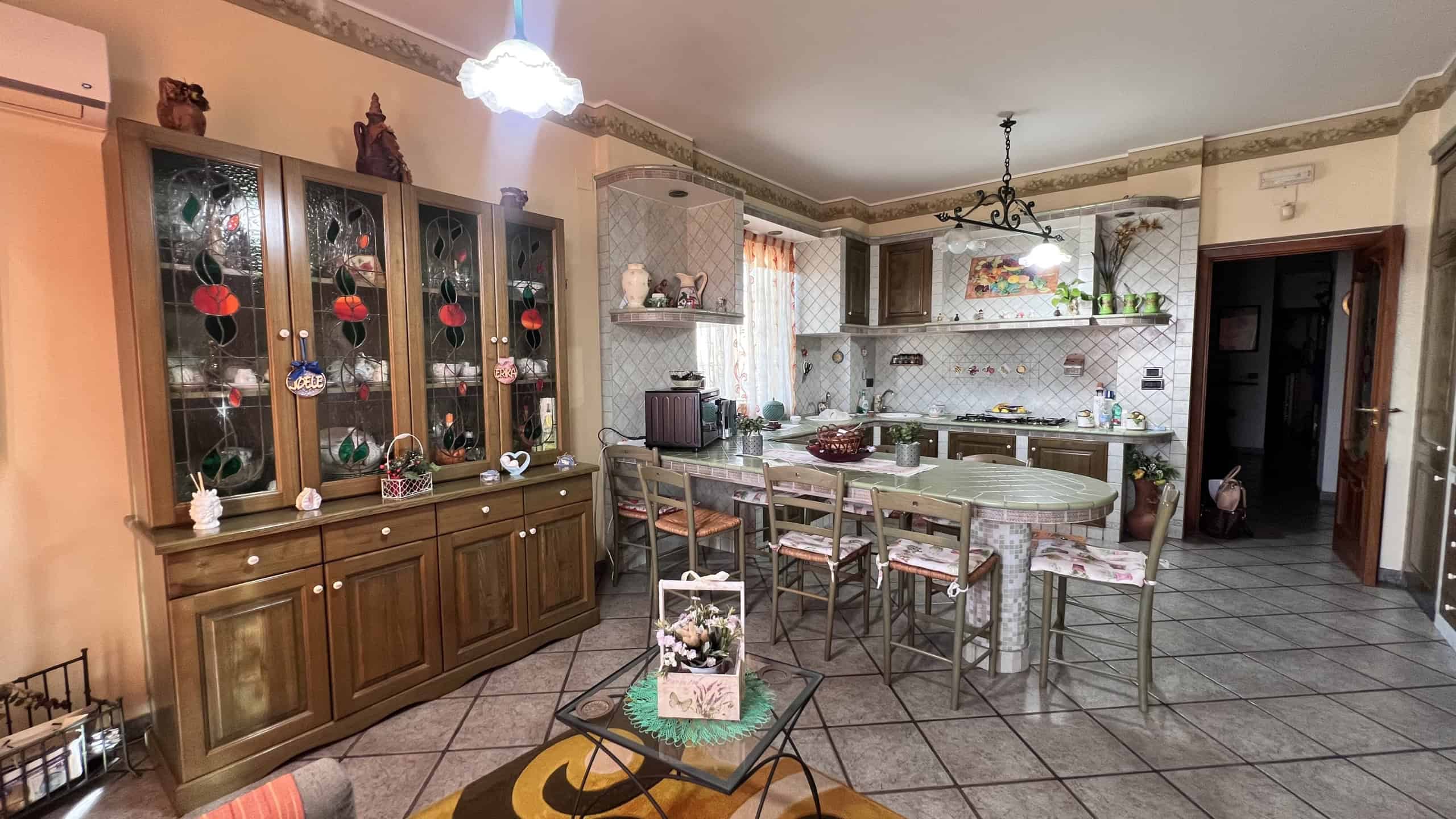 Interno di una cucina spaziosa e luminosa con mobili in legno, tavolo centrale e decorazioni floreali.