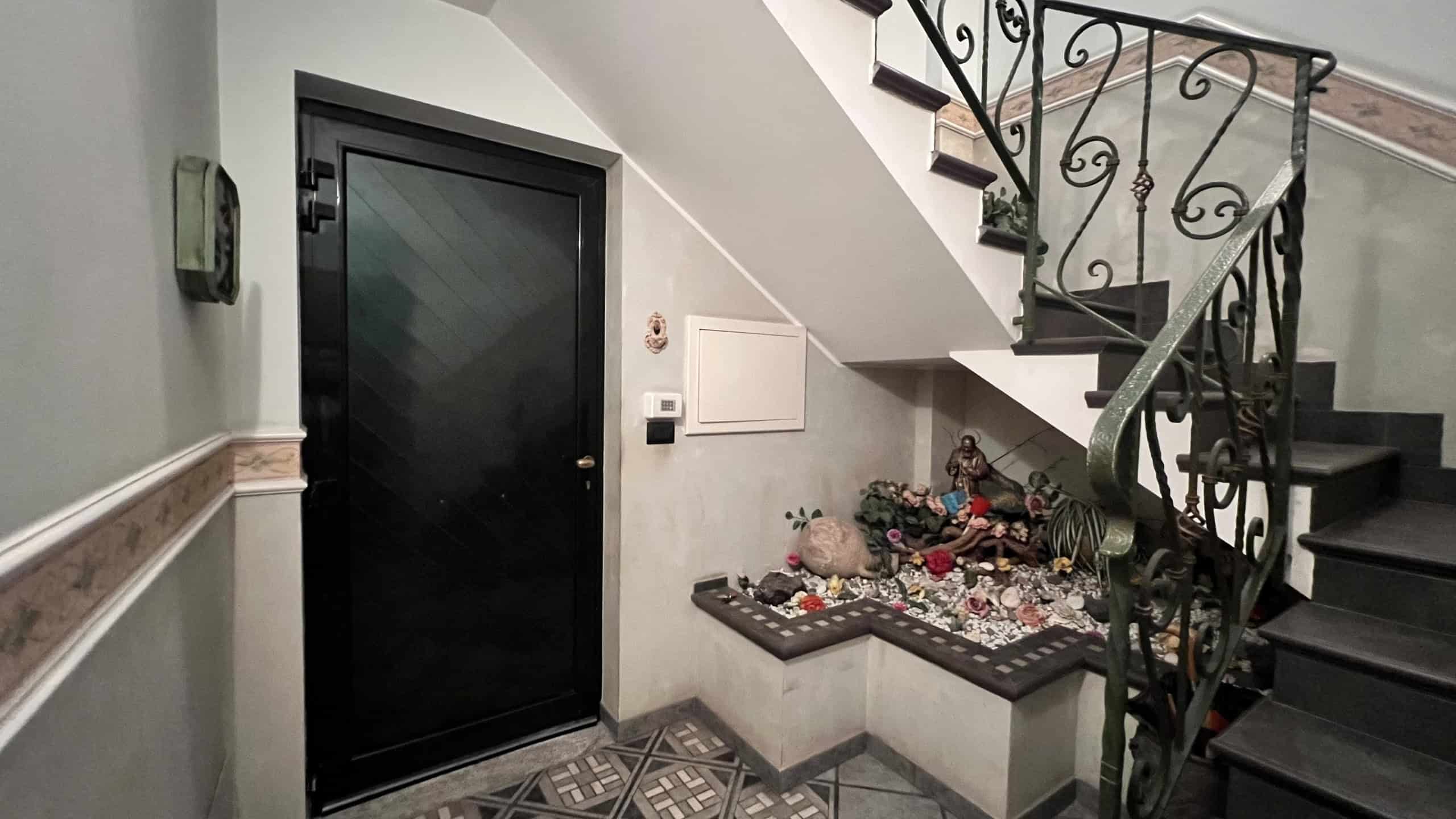 Ingresso di una villa unifamiliare con porta nera e scala in ferro battuto, decorazioni natalizie e oggetti vari sotto le scale.