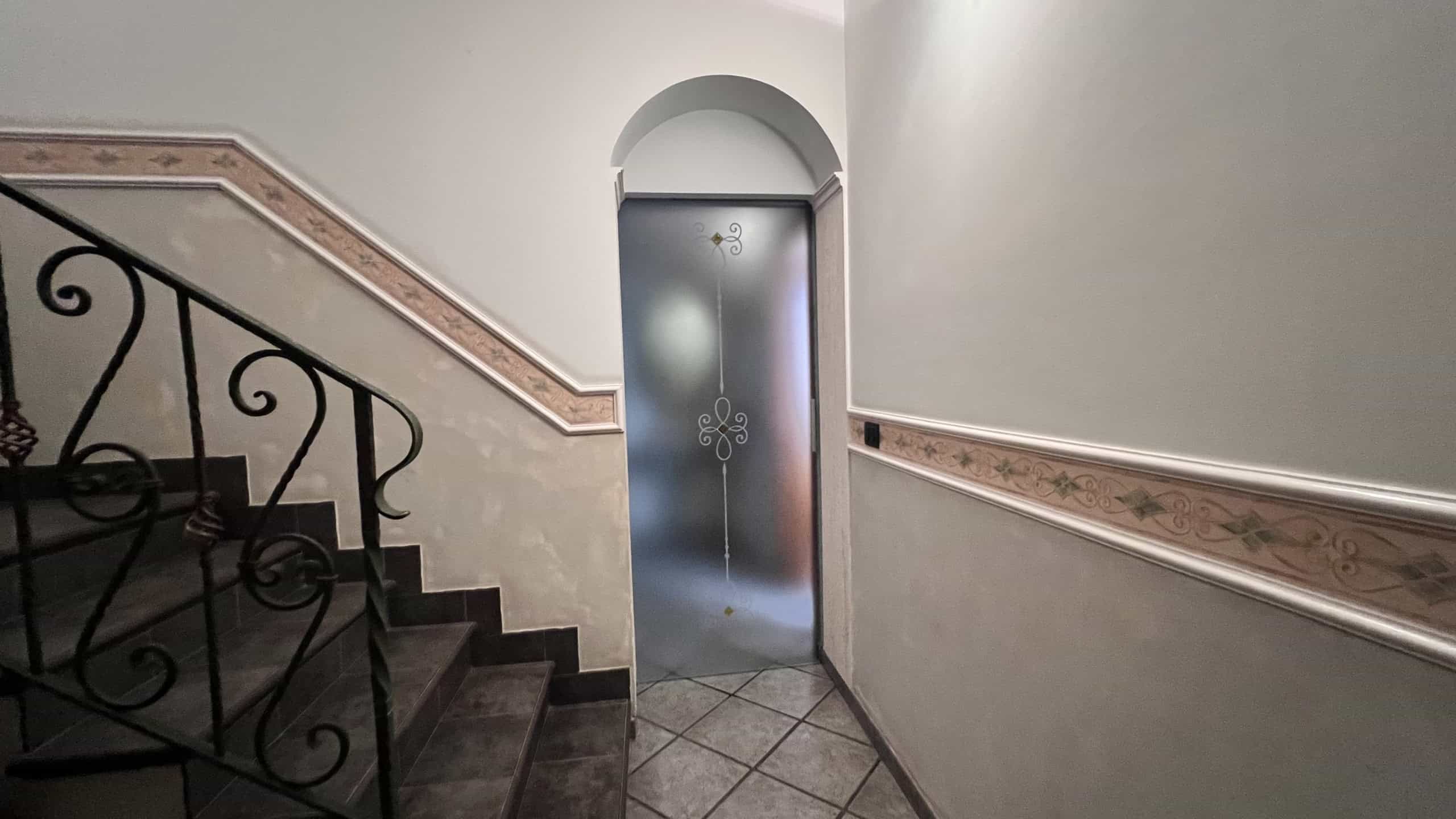 Ingresso condominiale con porta in vetro decorato e scale in marmo nero, ambiente luminoso e curato.