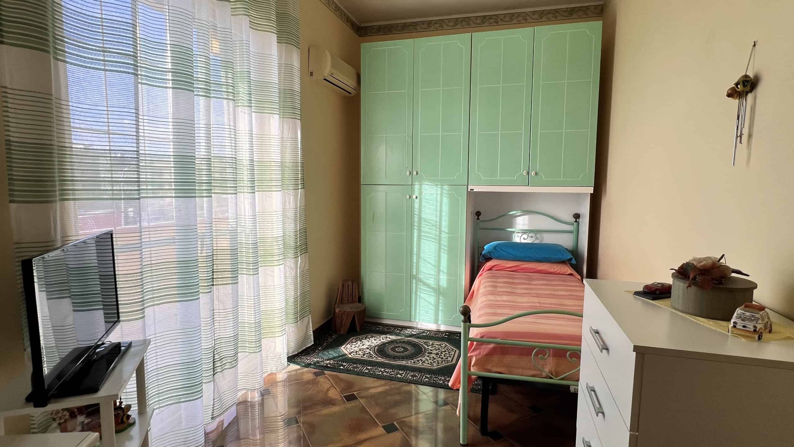 Camera da letto con armadio verde, letto singolo con copriletto rosa e finestra con tende leggere.