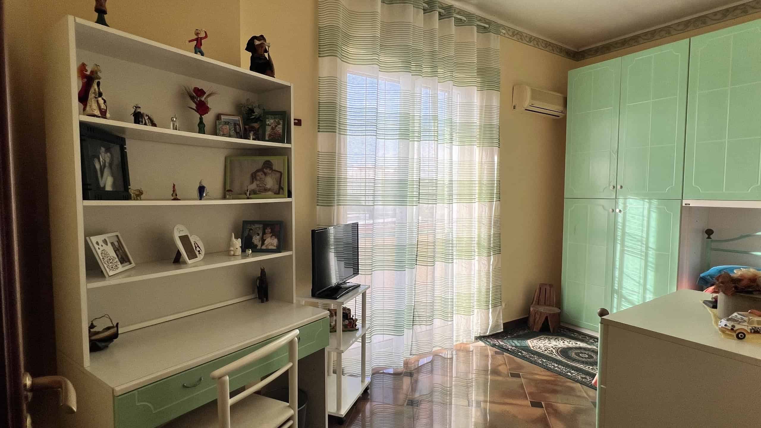 Interno di una casa con pareti color crema, armadio verde, libreria bianca e TV, con luce naturale da finestra con tende a righe.