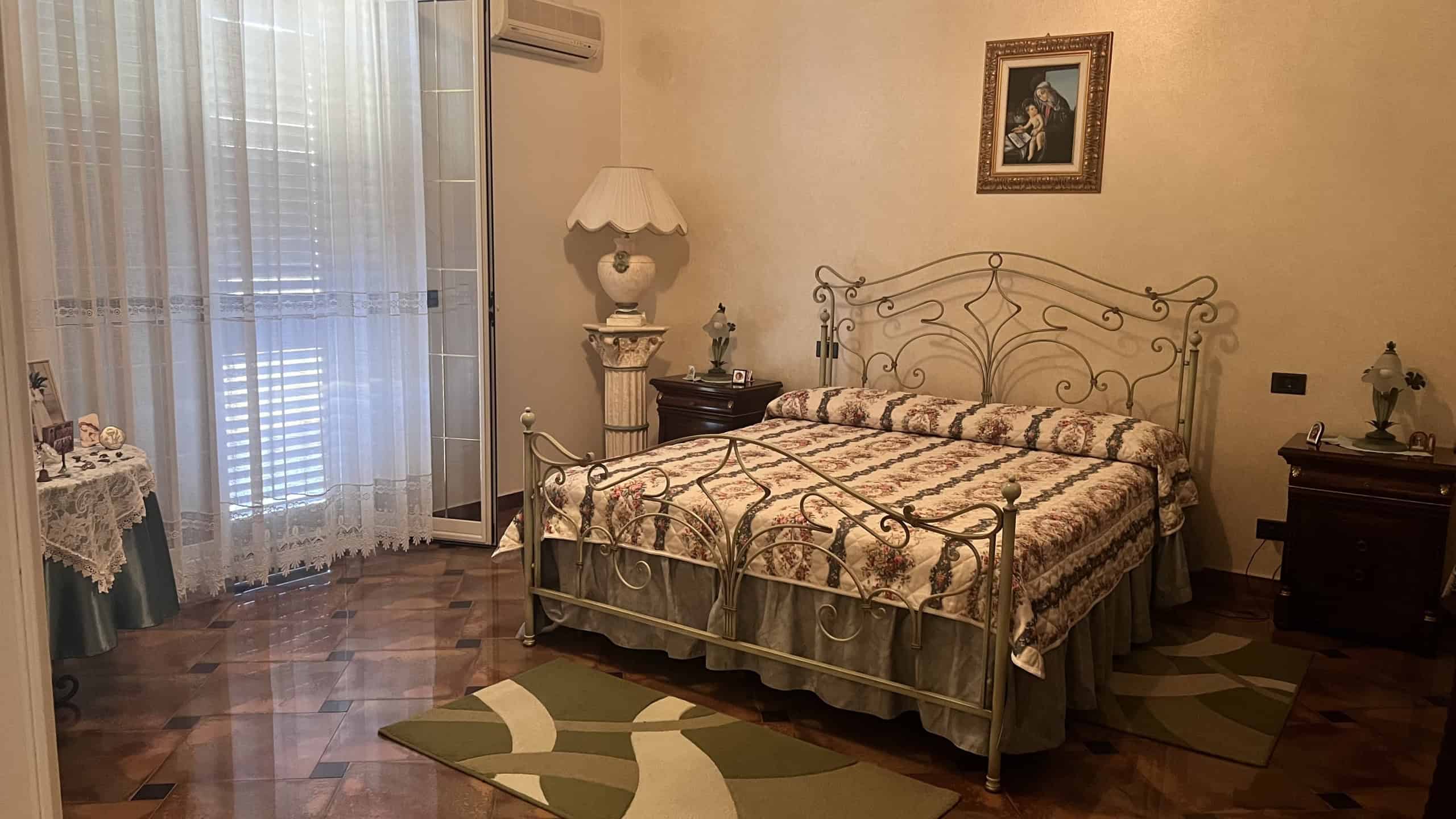 Camera da letto in stile classico con letto in ferro battuto, lampada da tavolo e mobili in legno, in un ambiente luminoso e accogliente.
