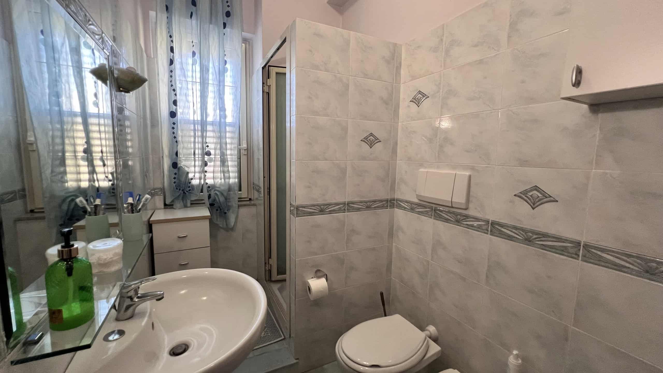 Bagno moderno con piastrelle chiare, lavabo, doccia e accessori essenziali.