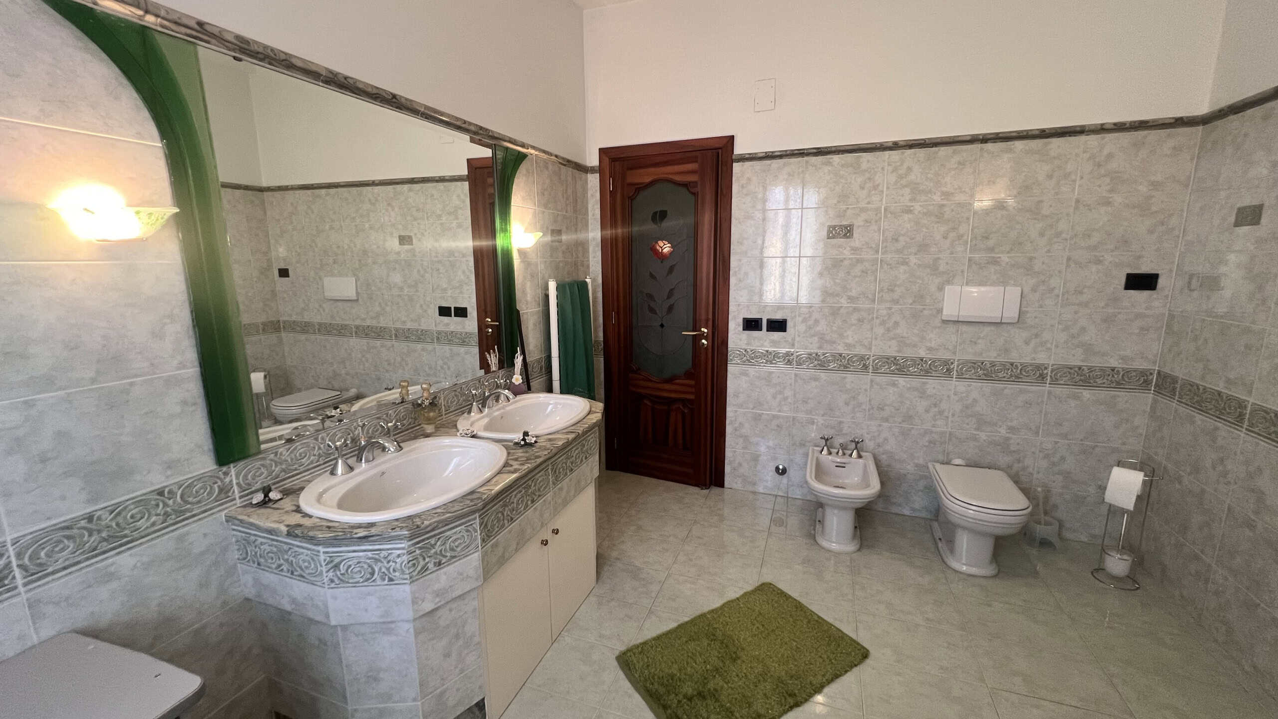 Bagno spazioso con doppio lavabo, bidet e WC, pareti rivestite in piastrelle grigie e specchio grande, ideale per una villa in vendita a Villafranca Tirrena.