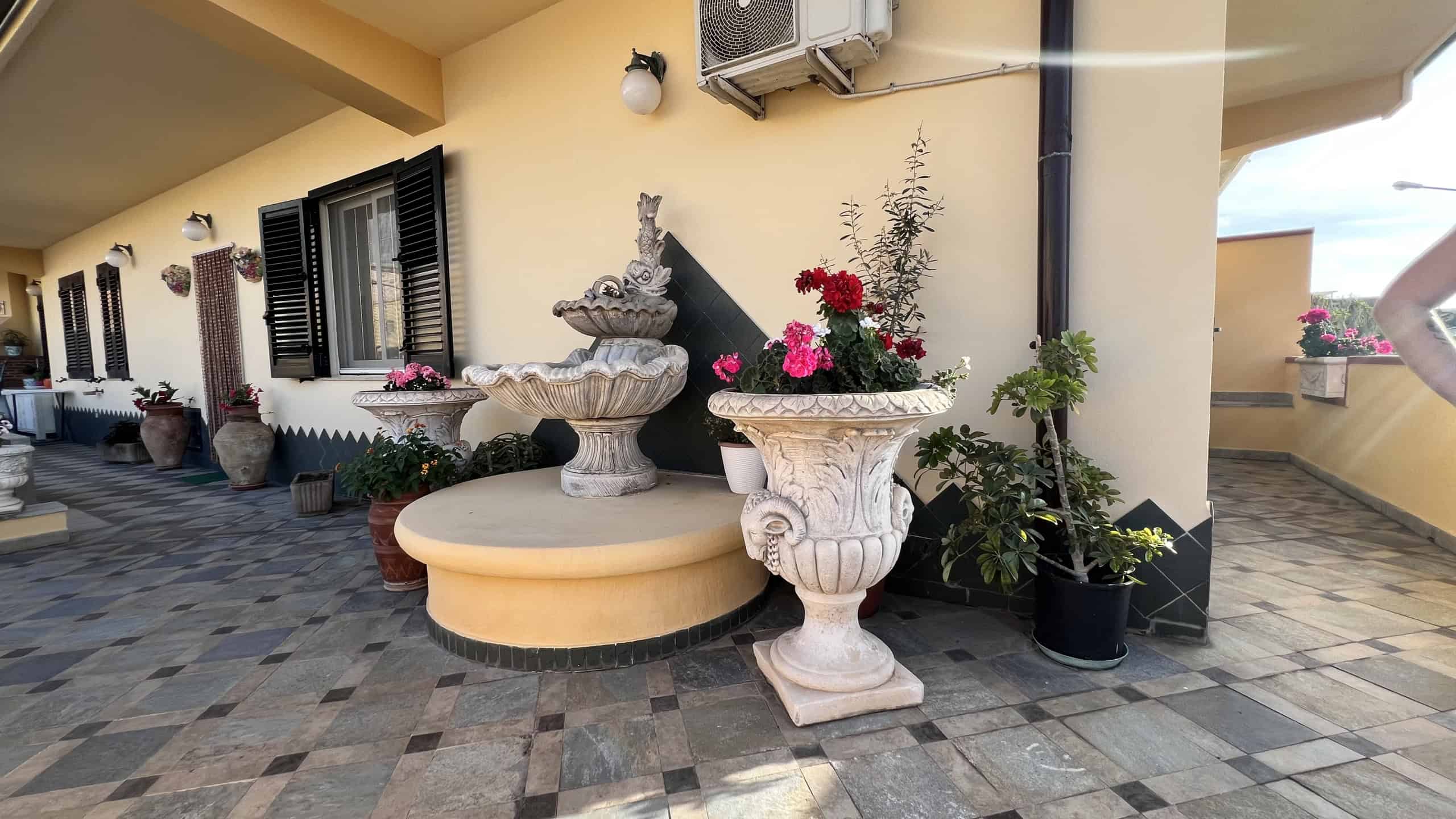 Balcone di una villa con fontana decorativa e piante fiorite, vista su un'area esterna ben curata a Villafranca Tirrena.
