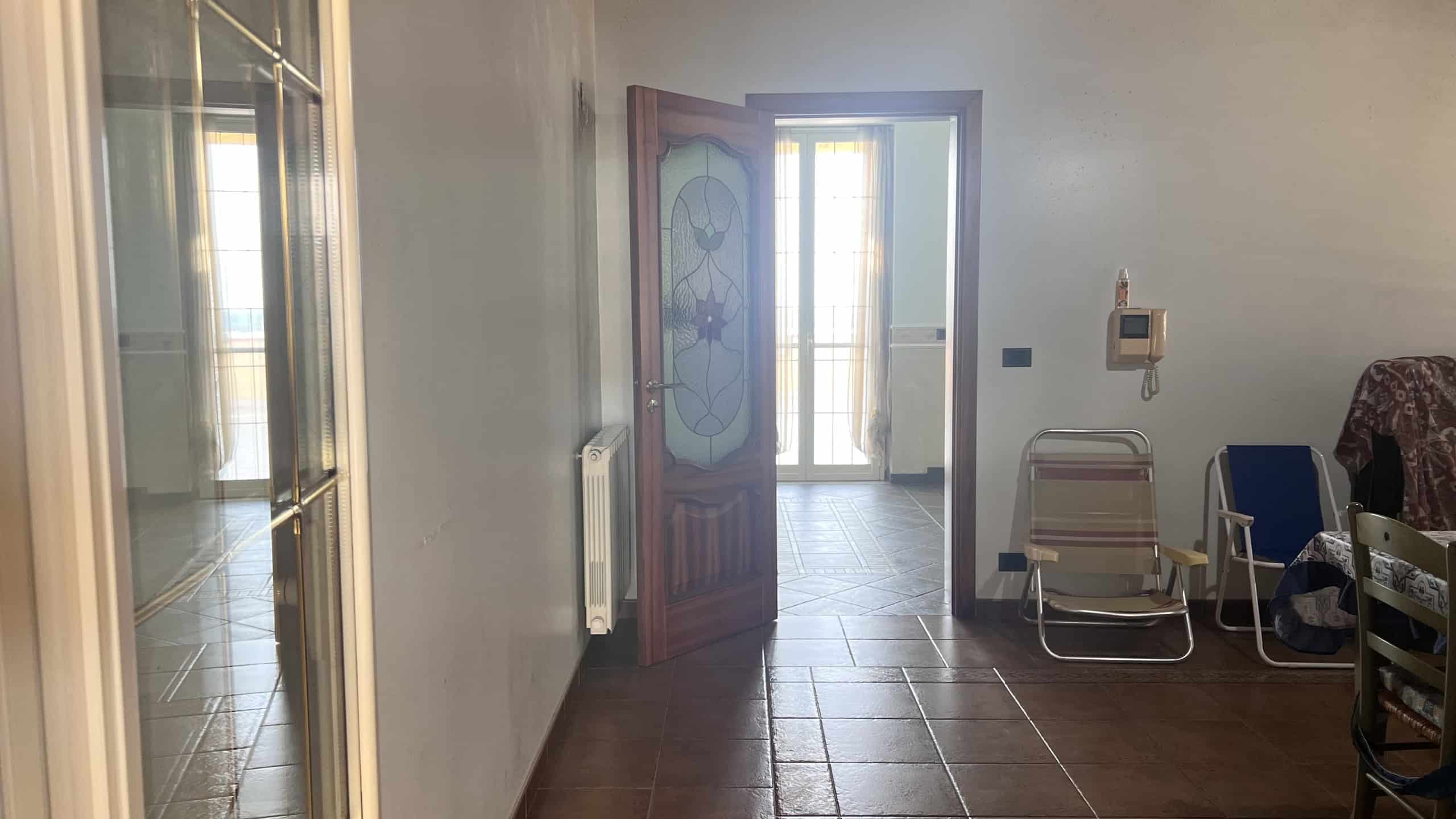 Ingresso di una villa unifamiliare con porta in legno e vetro decorato, vista su un soggiorno luminoso con balcone e mobili vari.