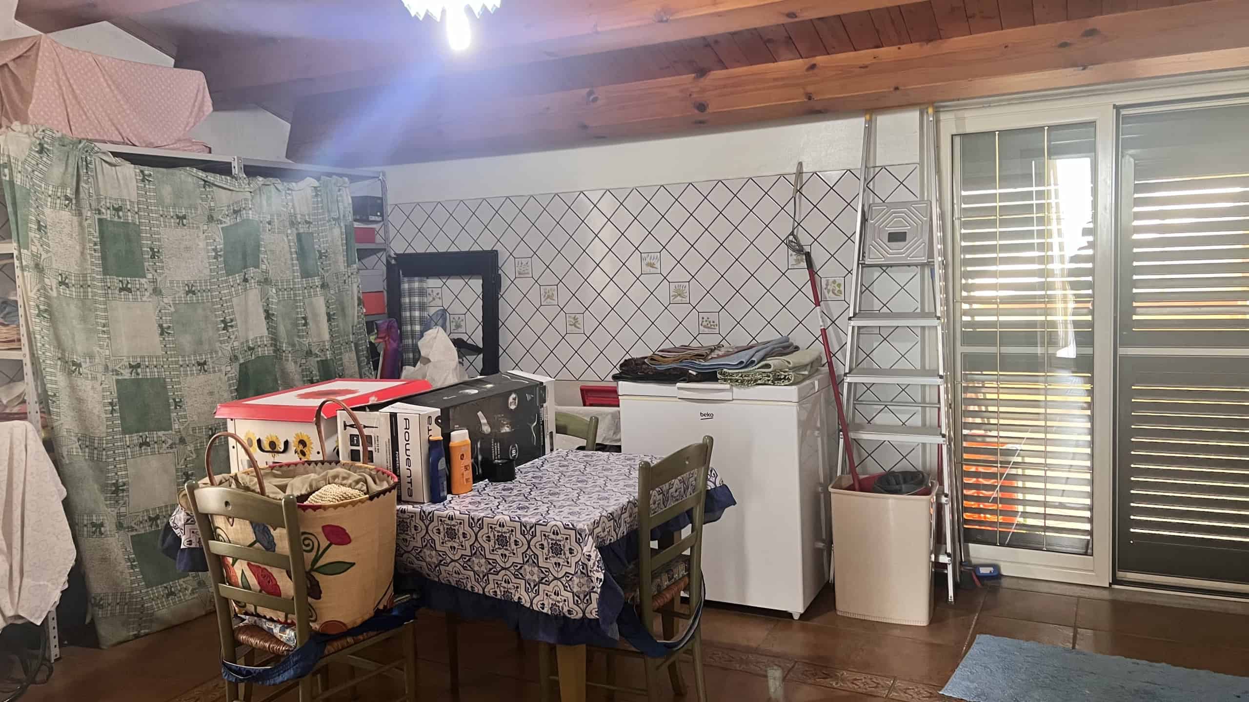 Interno di una casa con mobili, tavolo, sedie, e accessori vari, in un ambiente con pareti rivestite di piastrelle e soffitto in legno.