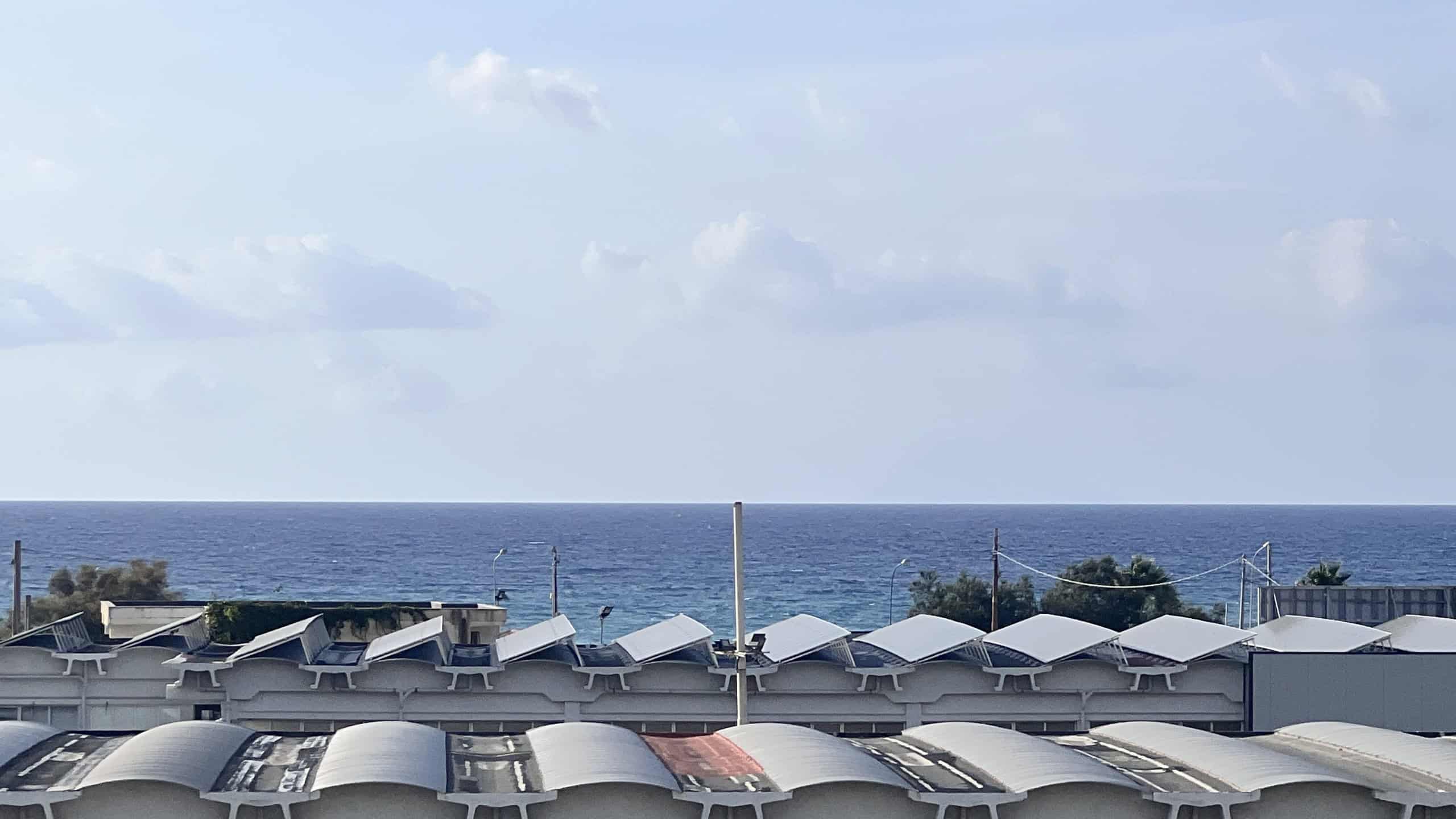 Vista panoramica del mare con case e strutture residenziali sullo sfondo, cielo sereno con nuvole sparse.