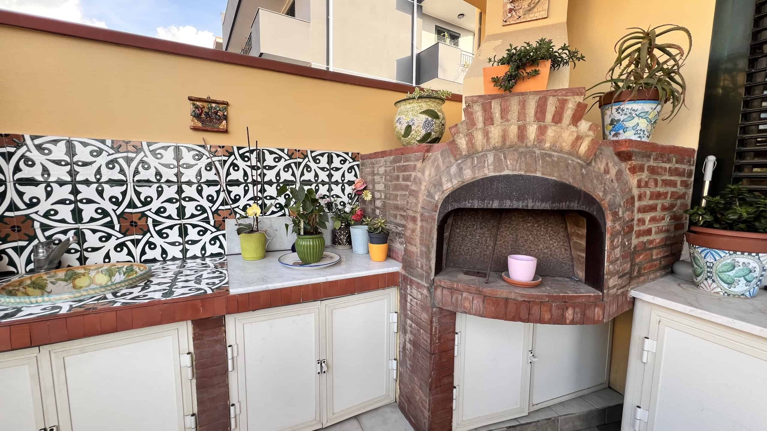 Angolo barbecue con forno in mattoni e piante decorative su terrazza a Villafranca Tirrena.