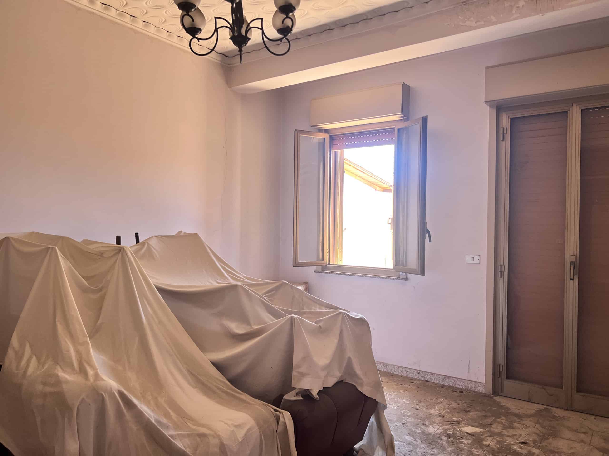 Vista di un soggiorno con divano coperto e finestra luminosa in un appartamento in vendita a Scaletta Zanclea, vicino a Messina.