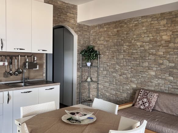 Interno di un soggiorno con cucina moderna, parete in pietra, tavolo con sedie e divano con cuscino decorativo.