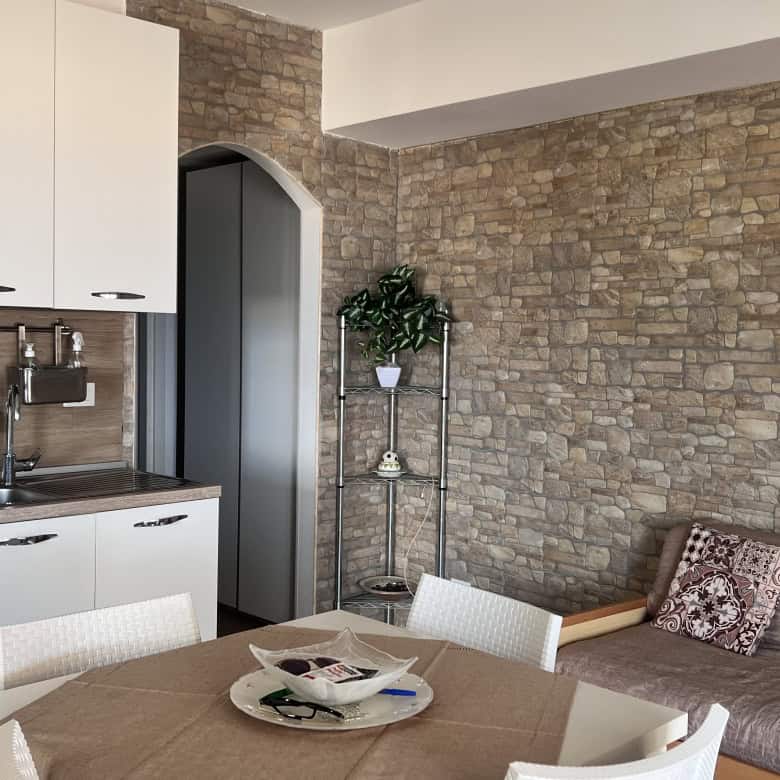 Interno di un soggiorno con cucina moderna, parete in pietra, tavolo con sedie e divano con cuscino decorativo.