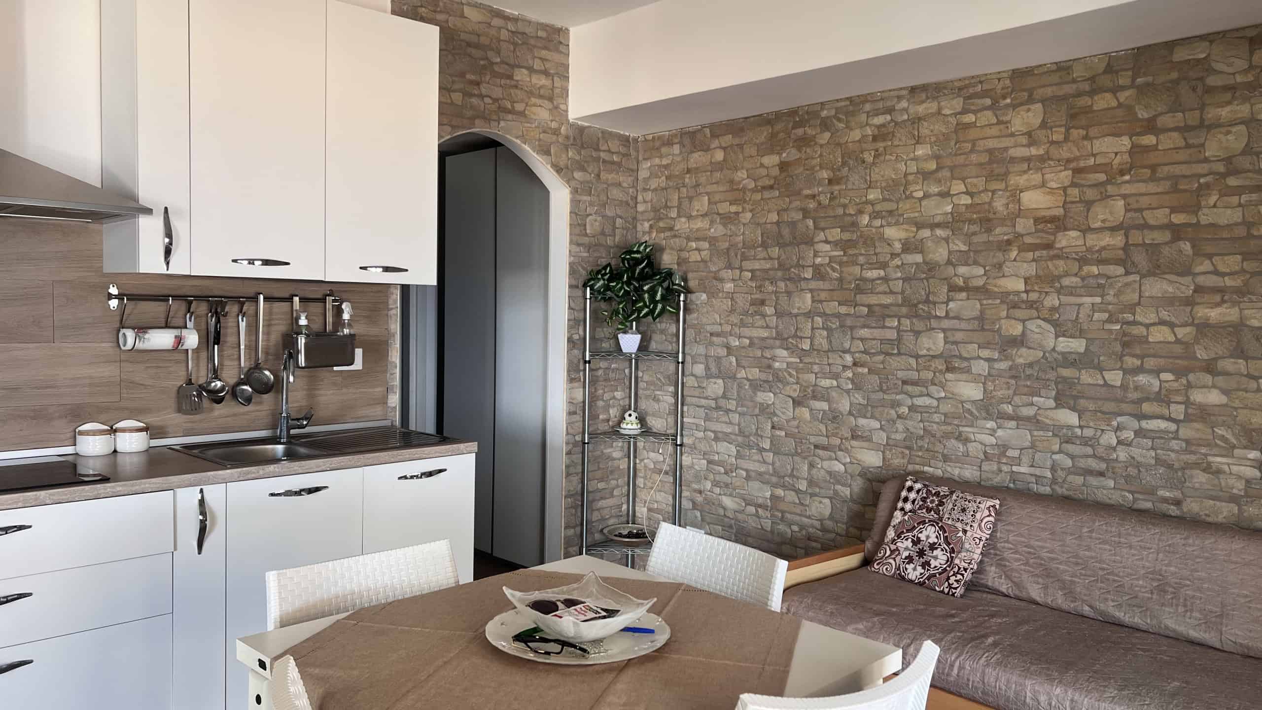 Interno di un soggiorno con cucina moderna, parete in pietra, tavolo con sedie e divano con cuscino decorativo.