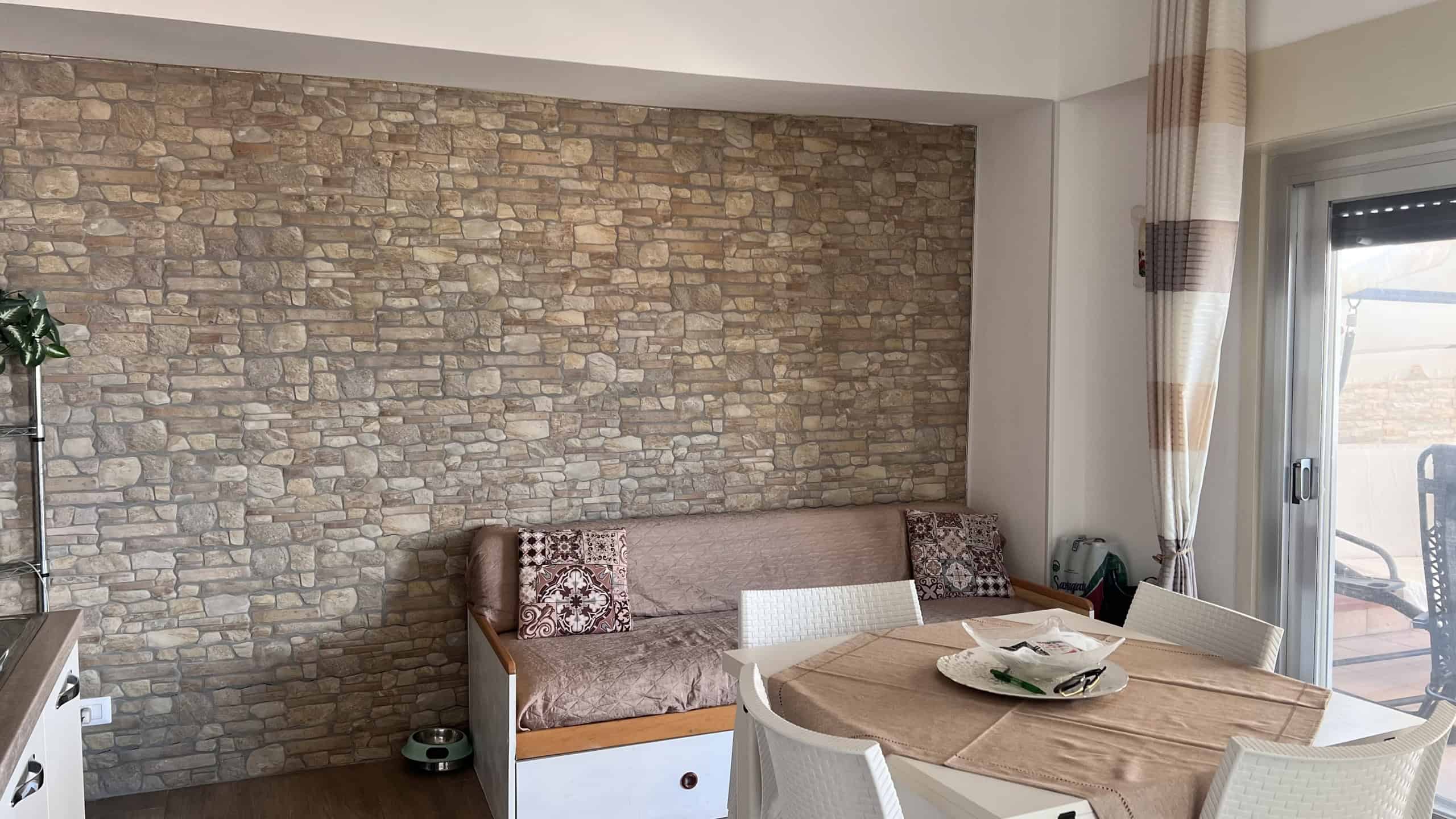 Interno di un soggiorno con parete in pietra, tavolo rotondo e divano con cuscini decorativi, balcone visibile dalla finestra.