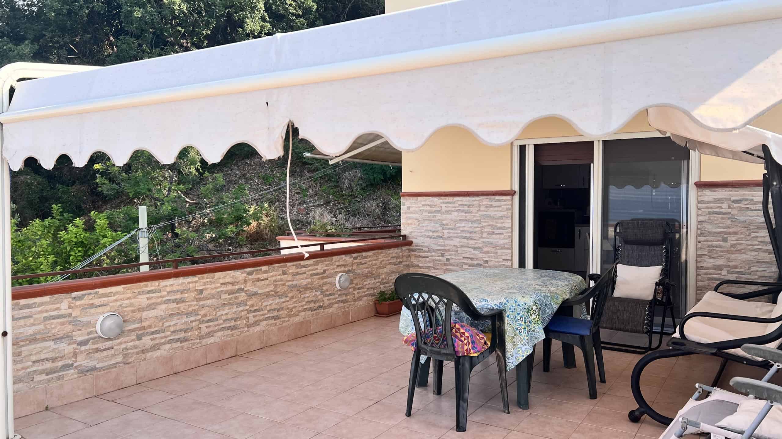 Terrazza spaziosa con tavolo, sedie e ombrellone, vista su giardino e paesaggio circostante, ideale per relax e convivialità all'aperto.