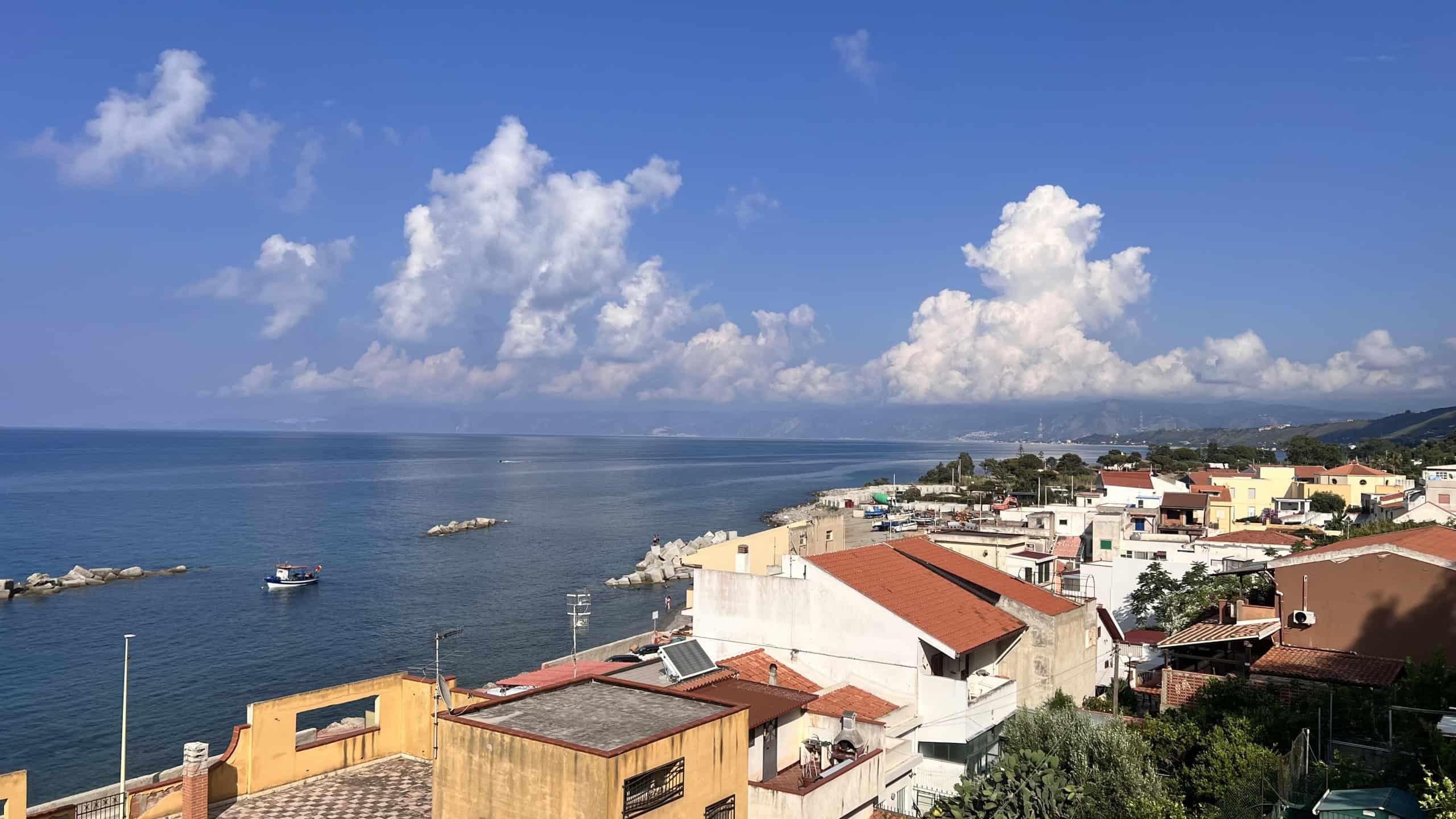 Panoramica vista mare con cielo sereno e nuvole bianche, vista dall'alto di Acqualadroni, Me, con case e porto sullo sfondo.