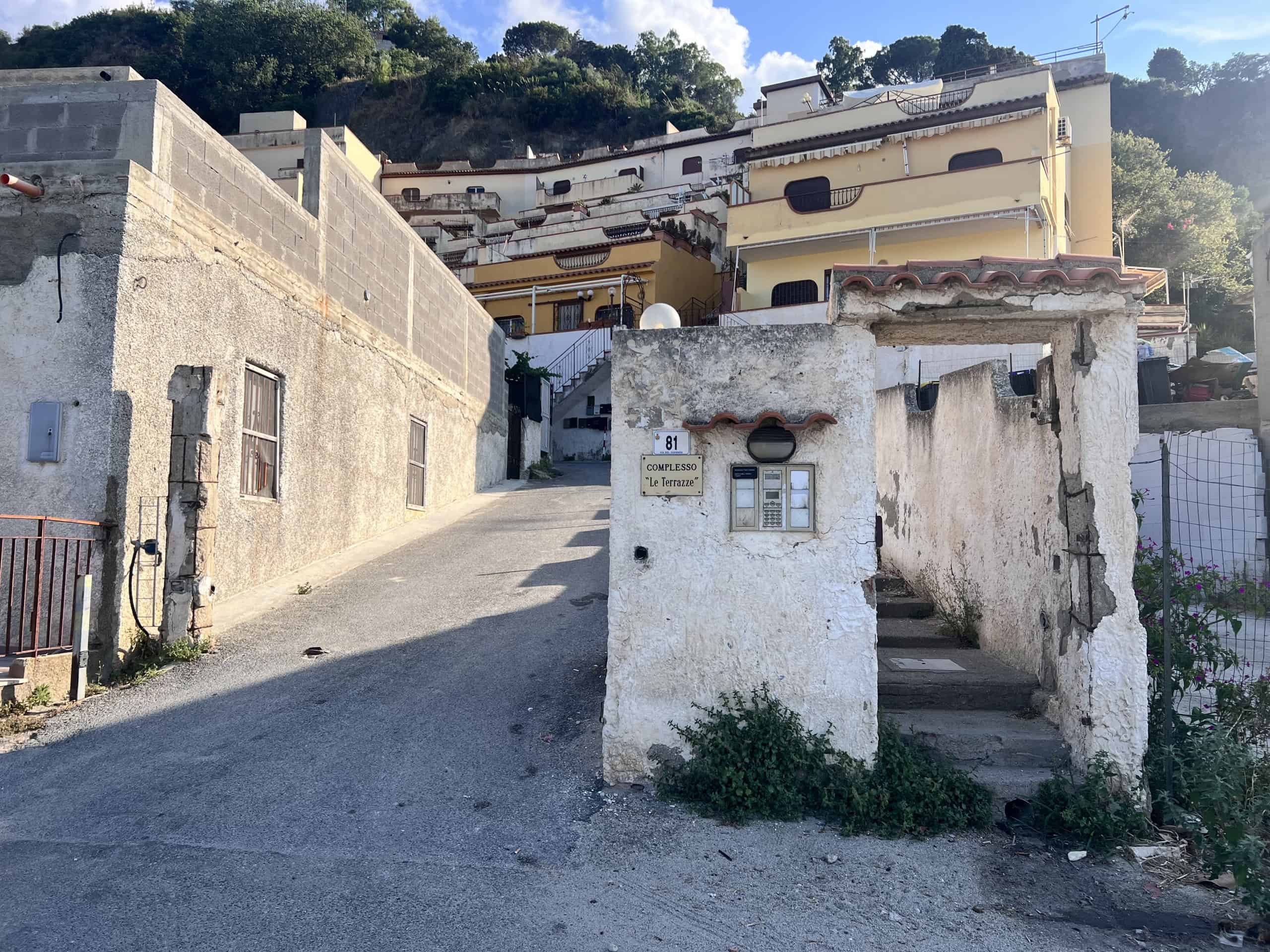 Vista di una strada in salita con case antiche e moderne a Acqualadroni, Me, in Sicilia, con un ambiente rurale e tranquillo.