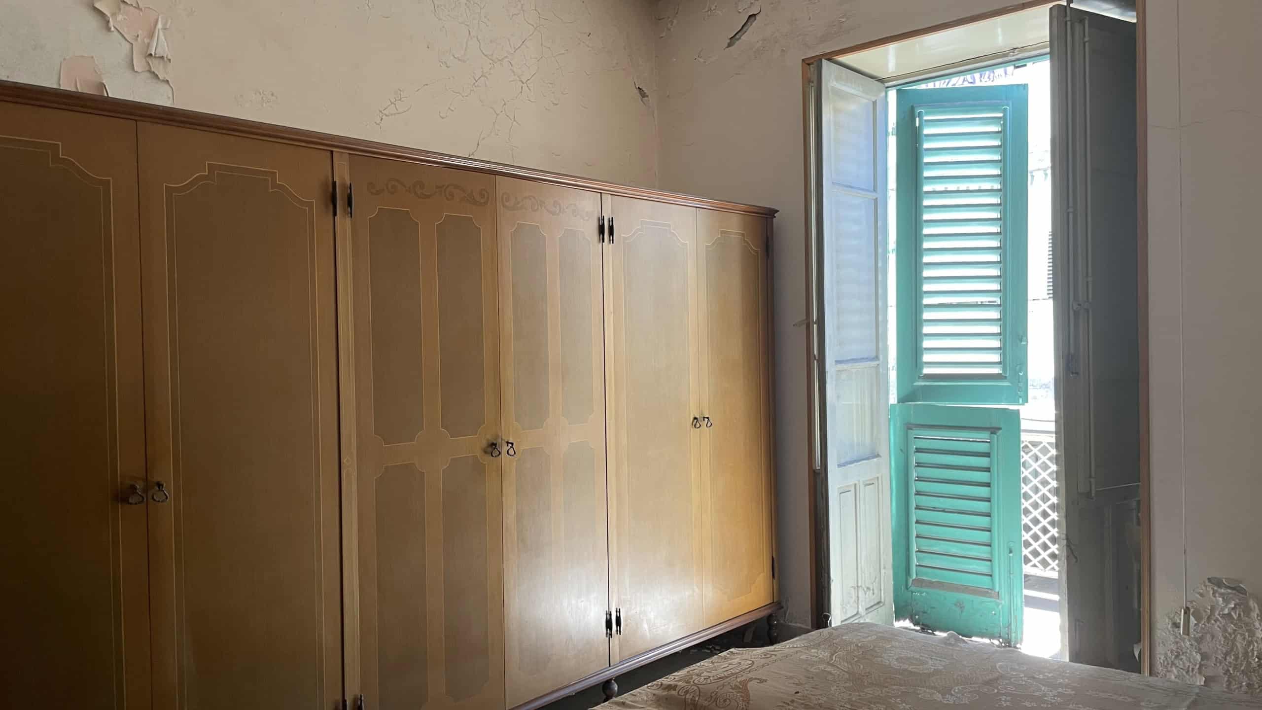 Camera da letto con armadio in legno e balcone con persiane verdi, luce naturale, pareti con segni di usura, pavimento in pietra.