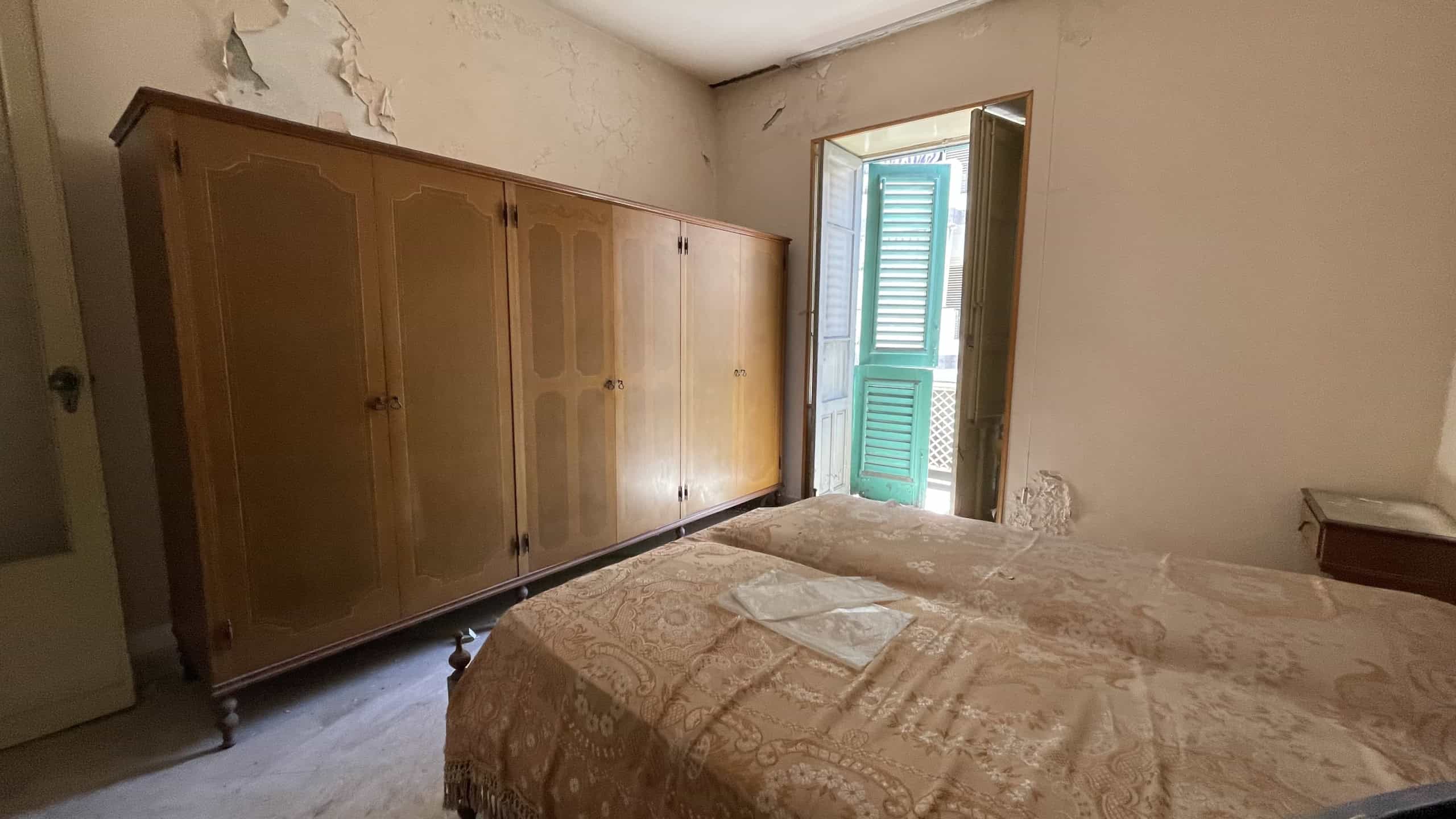 Camera da letto con armadio grande e porta finestra con vista esterna, ambiente datato e in condizioni di bisogno di ristrutturazione.