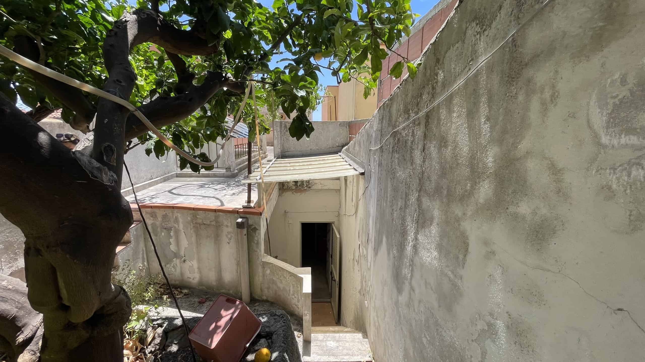 Vista di un cortile con albero, pareti in cemento e ingresso sotterraneo, in un contesto residenziale a Scaletta Zanclea, vicino a Via Roma 168.