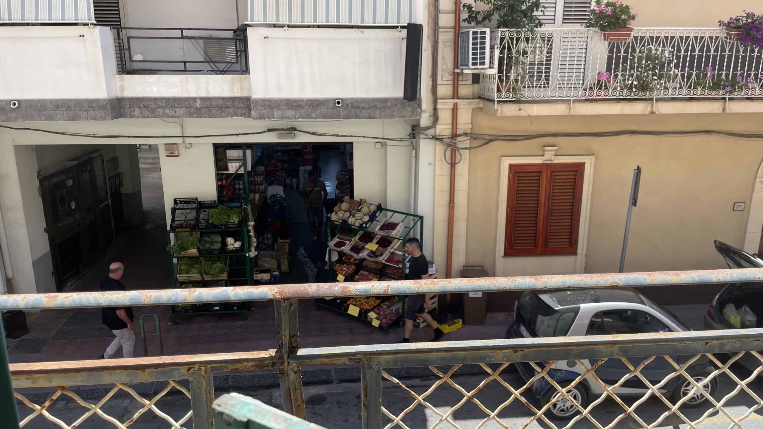 Venditore di frutta e verdura al mercato di Via Roma 168, Scaletta Zanclea, Me, con clienti e negozio in background.