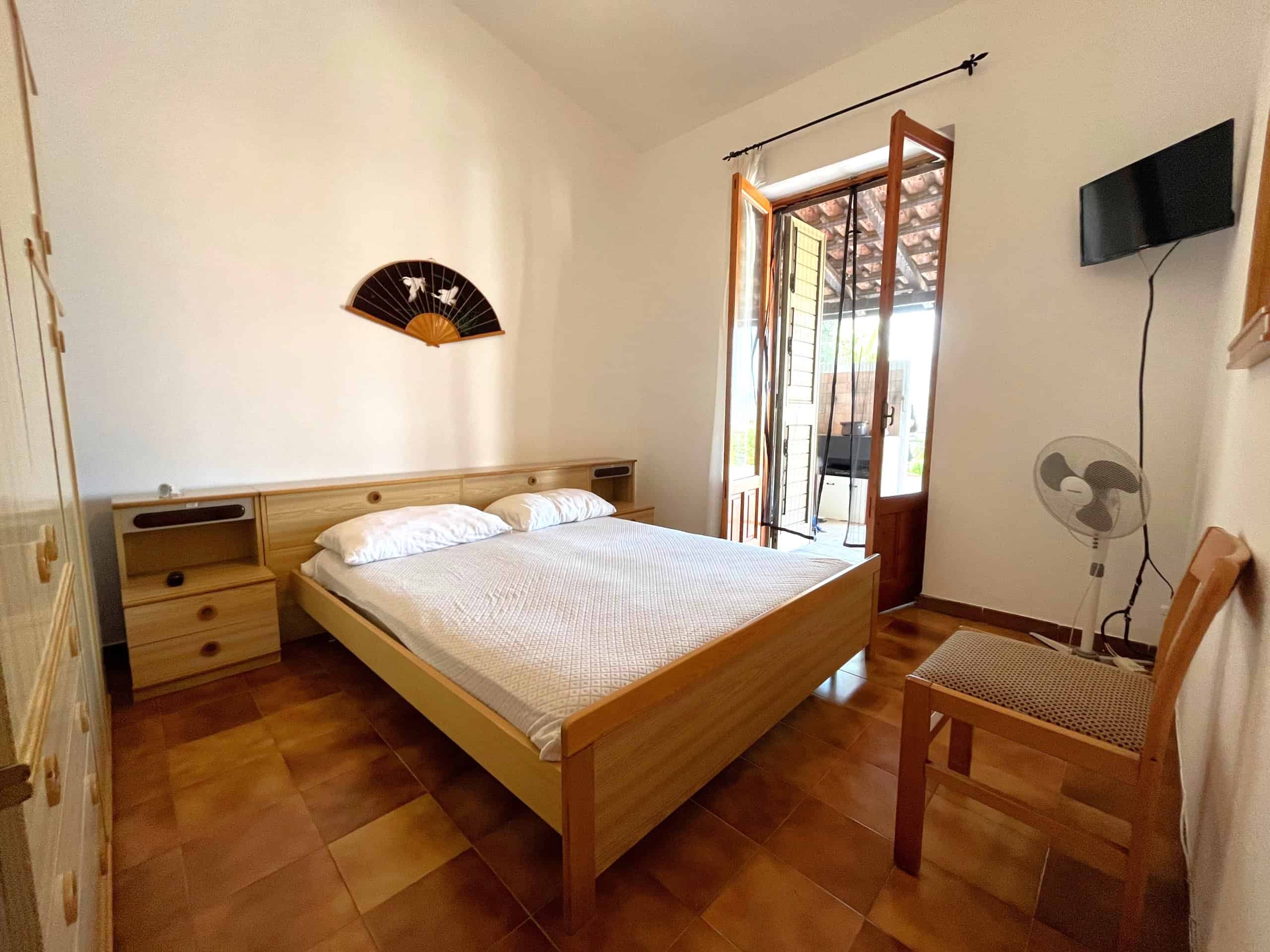 Camera da letto luminosa con letto matrimoniale, armadio, sedia, ventilatore e TV, con porta finestra che dà su un balcone.