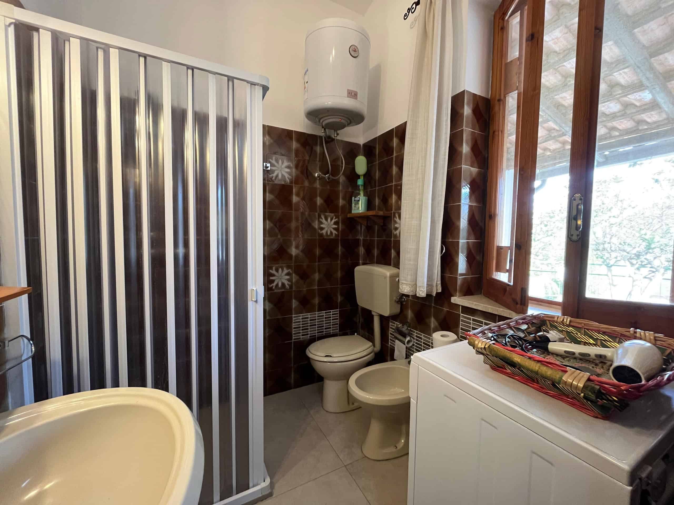 Bagno di una villetta in affitto con doccia, WC e lavatrice, finestra con tende, pareti rivestite in piastrelle marroni.