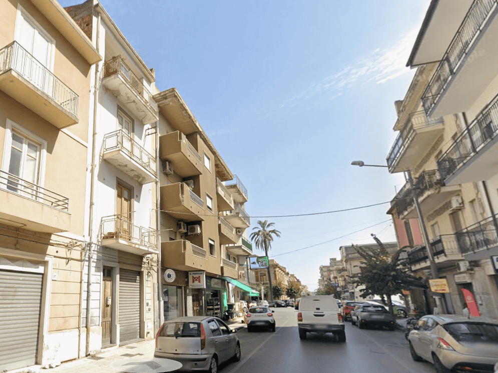Vista di una strada urbana con edifici residenziali e negozi, traffico veicolare e cielo sereno, in un quartiere di città italiana.