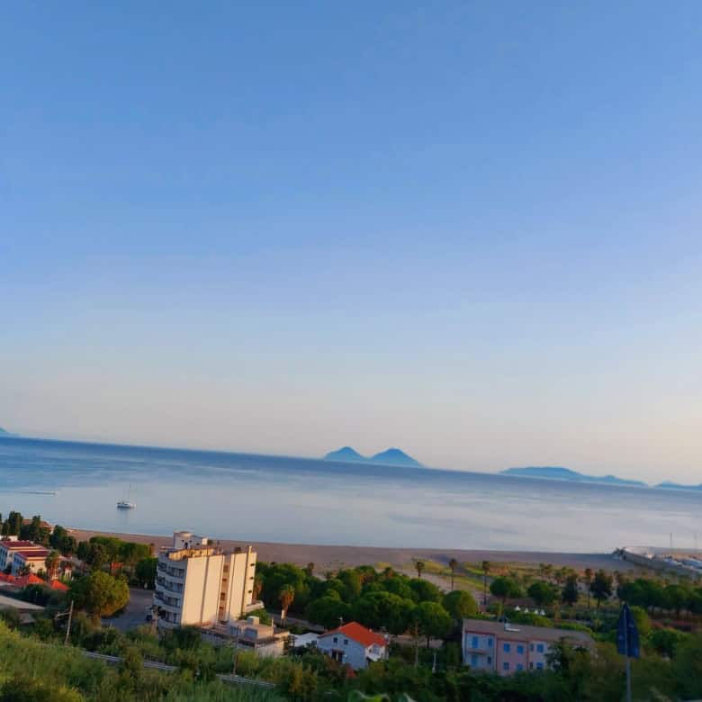 Vista panoramica del mare con isole sullo sfondo, case e vegetazione in primo piano, cielo sereno, rappresentando un paesaggio costiero tranquillo e suggestivo.