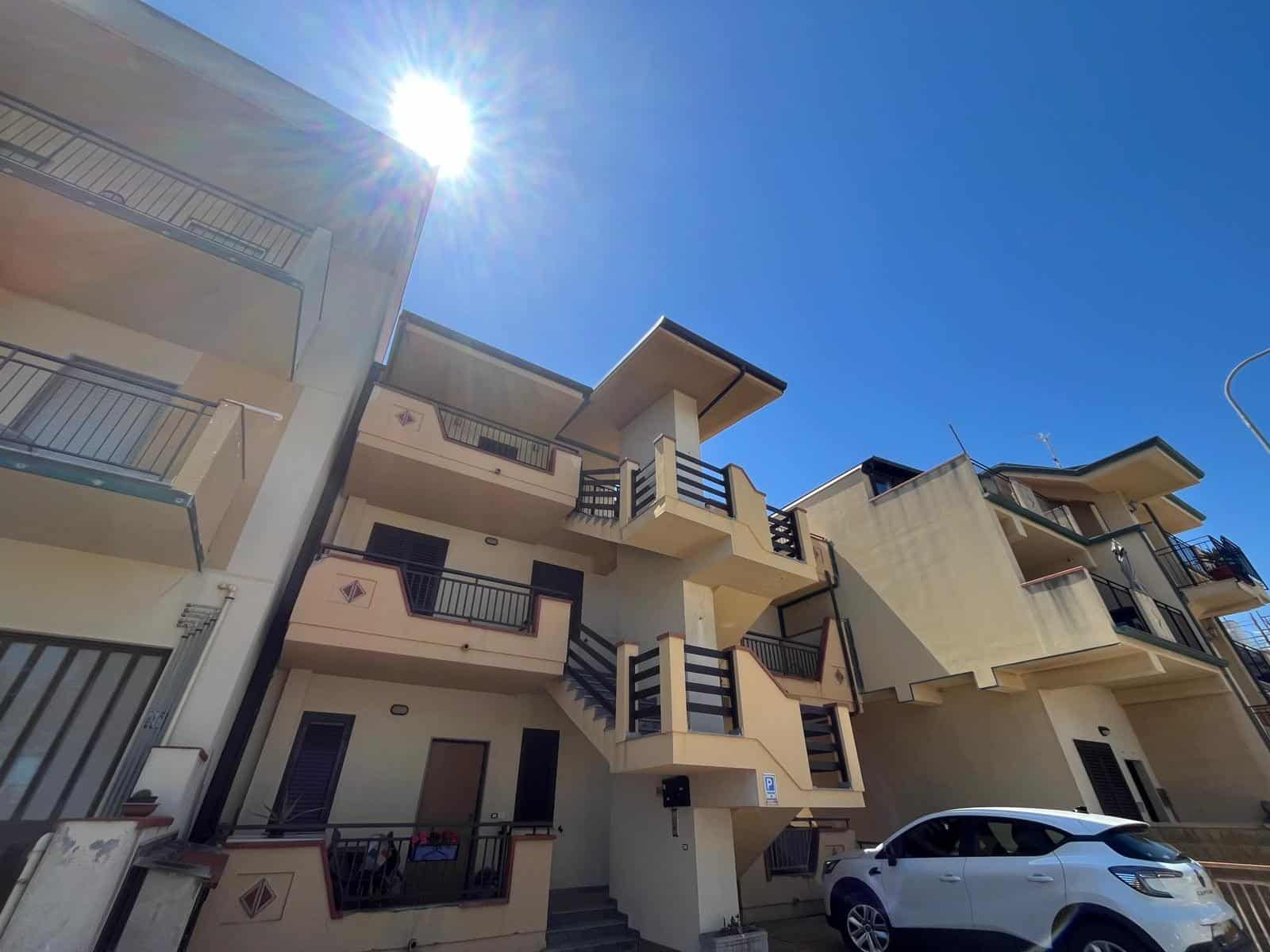 Ampia vista di un edificio residenziale moderno con balconi e terrazze, sotto un cielo sereno e soleggiato, con un'auto parcheggiata nel cortile. Ideale per immobili di nuova costruzione o appartament.