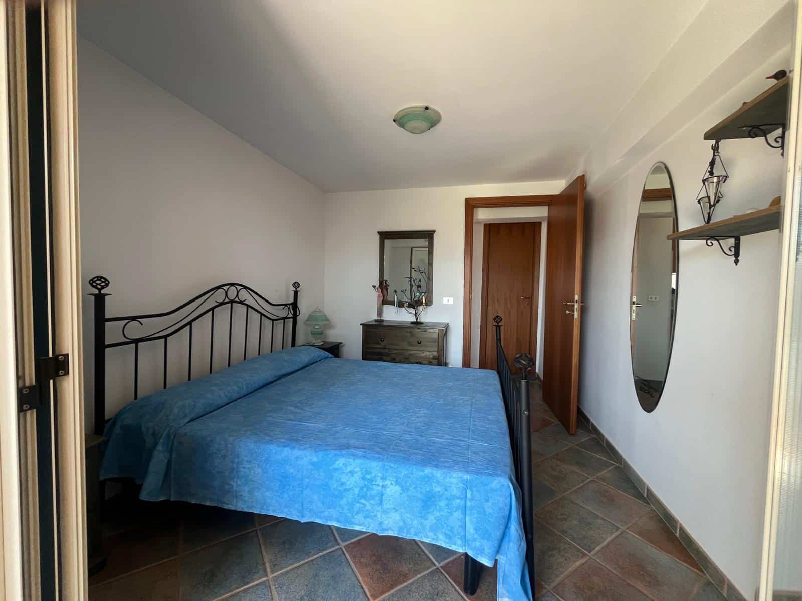 Il letto matrimoniale con copriletto blu in una camera da letto luminosa e accogliente, con pareti bianche, specchio rotondo e arredi in stile semplice e funzionale, ideale per una casa moderna o rist.