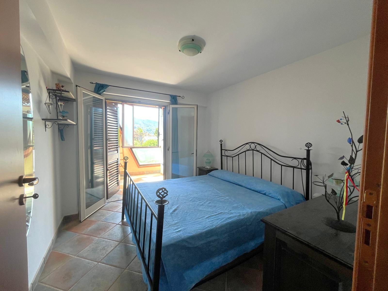 Letto matrimoniale con testiera in ferro battuto in una camera luminosa con balcone e vista sul cielo e gli alberi.
