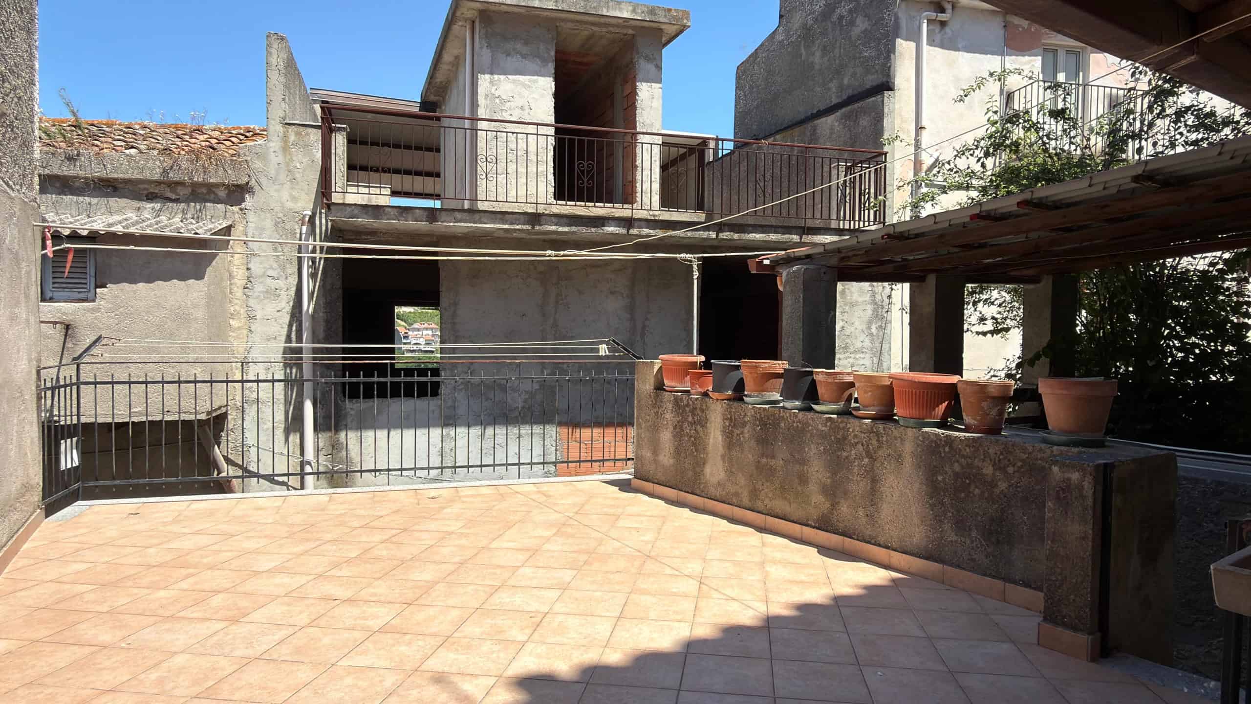 Balcone di un edificio in costruzione con piante in vaso e vista su un cortile esterno, in fase di completamento, con struttura in cemento e ringhiera in metallo.