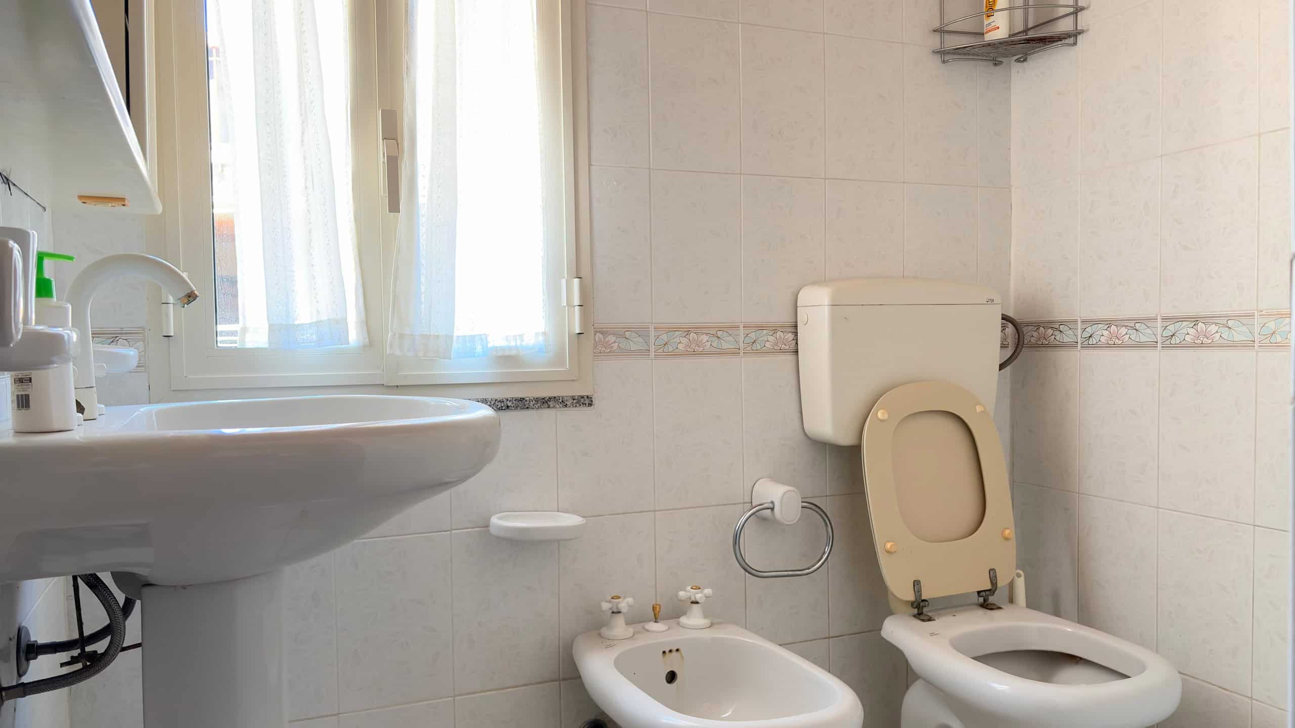 Lavandino e WC in un bagno moderno con pareti rivestite di piastrelle chiare, finestra con tende leggere, accessori per il bagno e illuminazione naturale, ideale per ambienti di comfort e funzionalità.