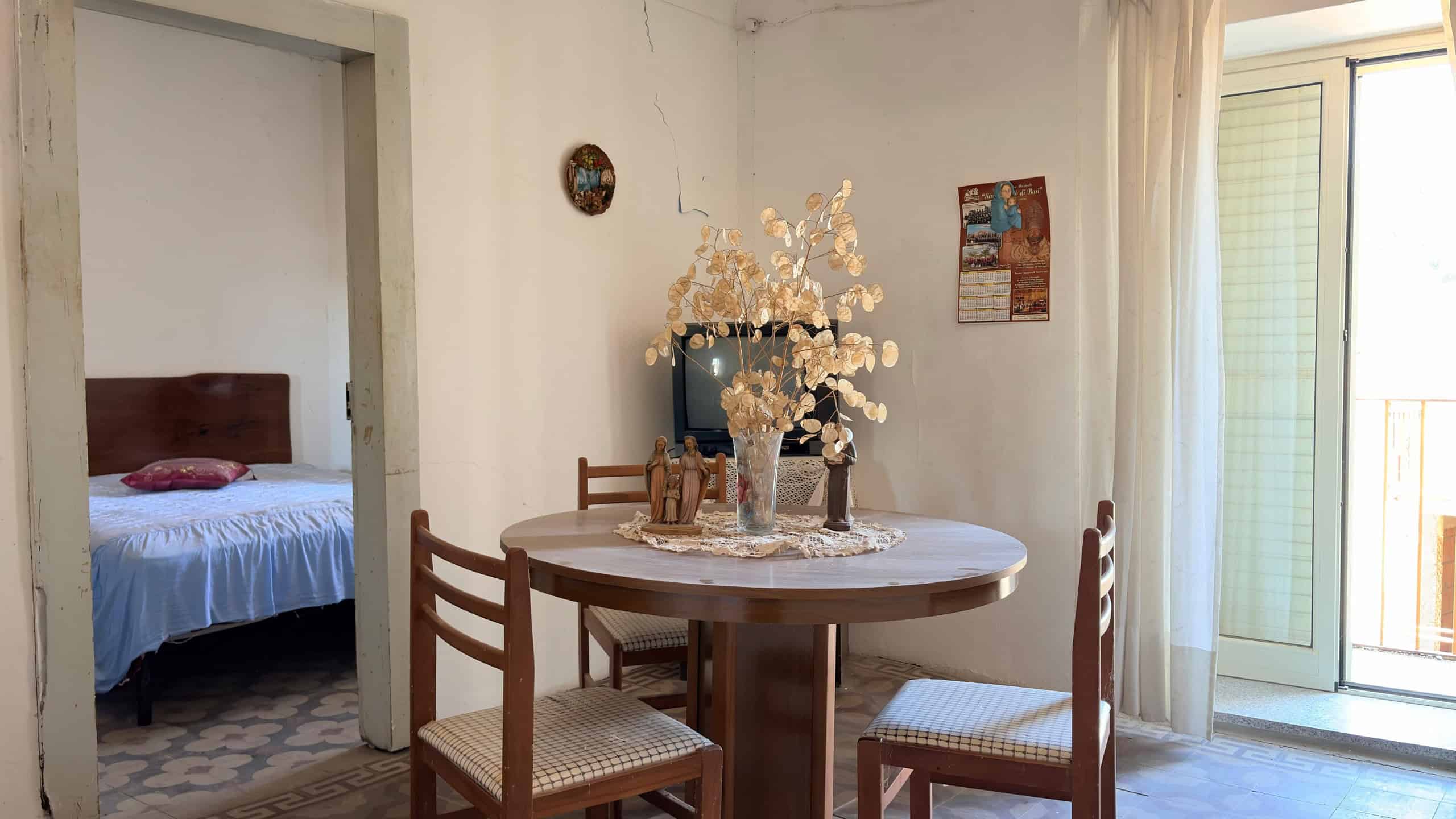 Elegante soggiorno con tavolo rotondo in legno, decorato con fiori freschi e accessori raffinati, illuminato da luce naturale proveniente da una porta finestra con tende leggere.